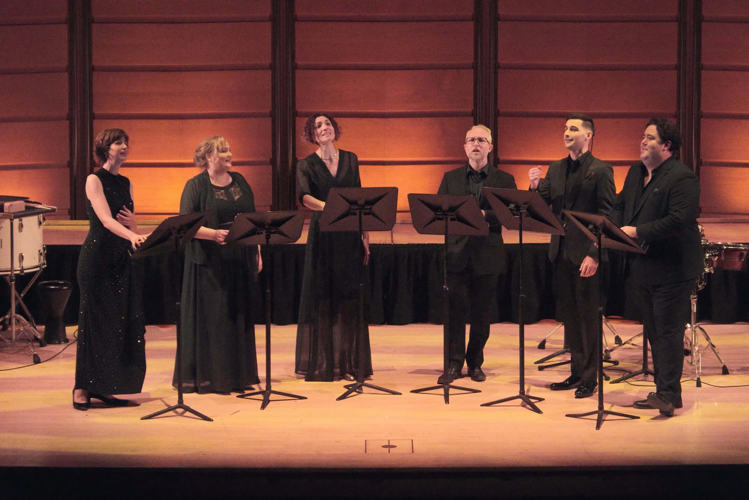 A group of seven classical singers performing on a stage with music stands, in front of a warm, wooden-paneled background, dressed in formal black attire.