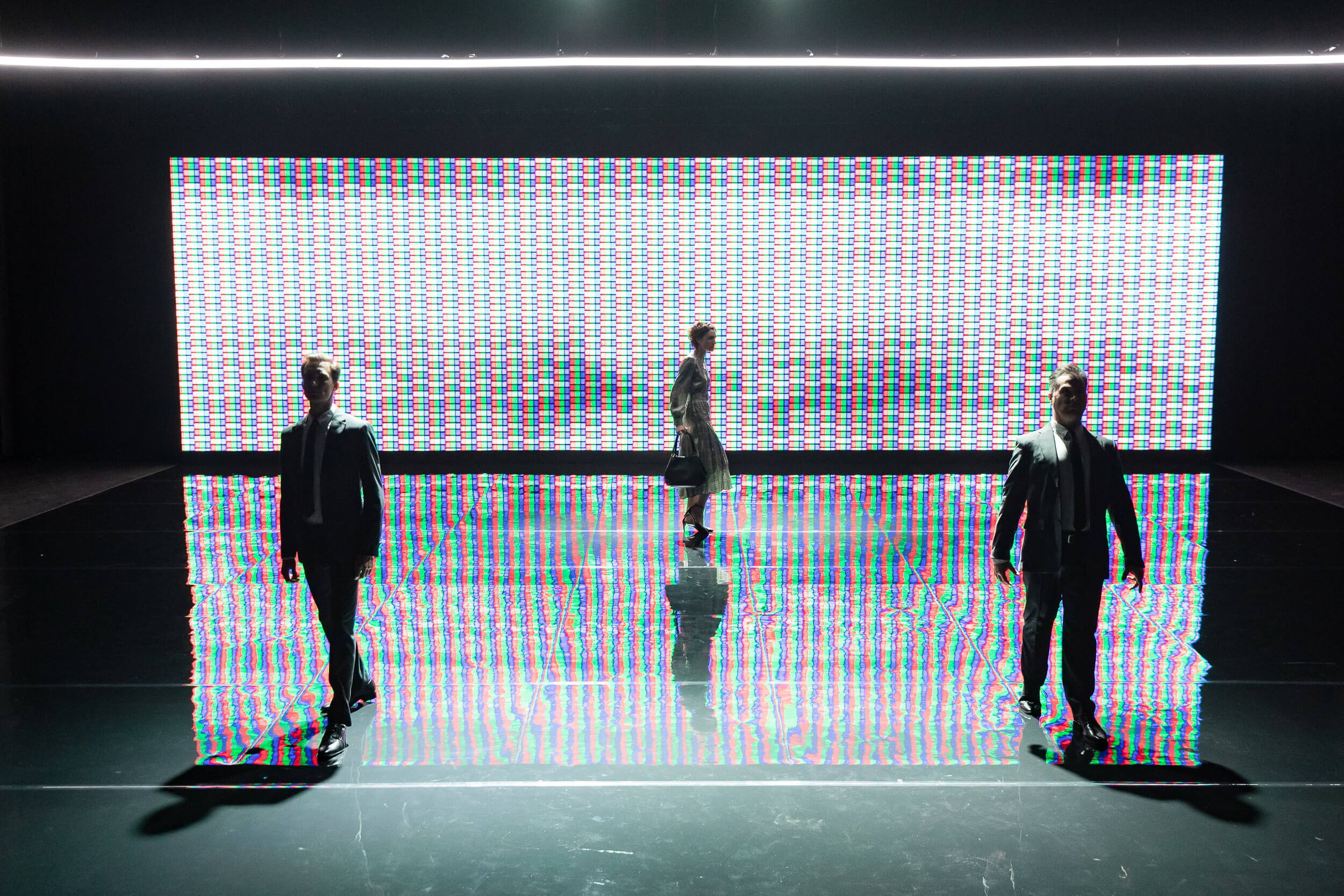 Four people in formal attire walk in front of a large digital screen displaying a colorful pixelated pattern, with the reflections visible on a shiny floor.