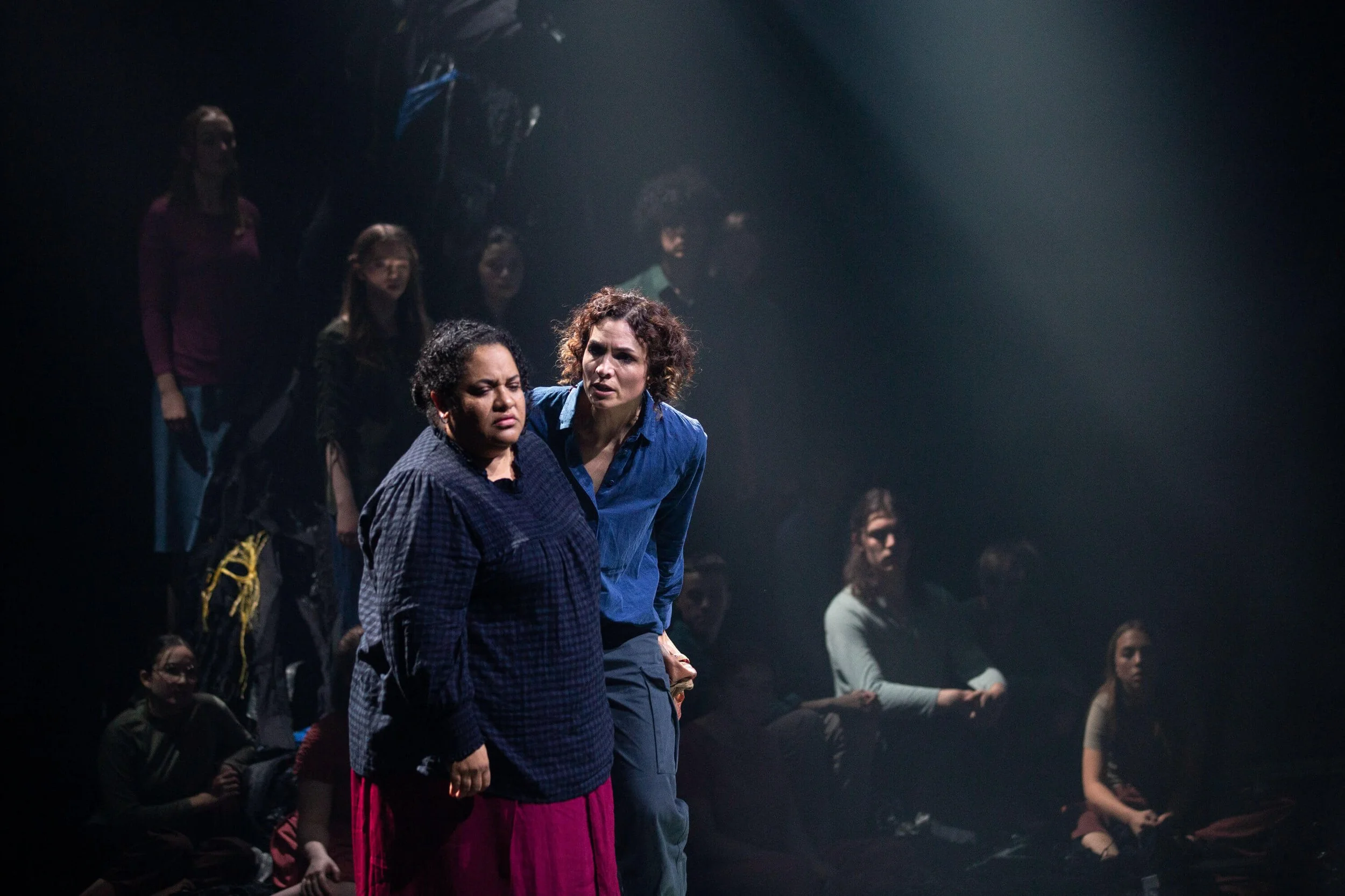 People standing closely together in a dark setting, with some illuminated by spotlight, indicating a serious or emotional moment during a theatrical performance or event.