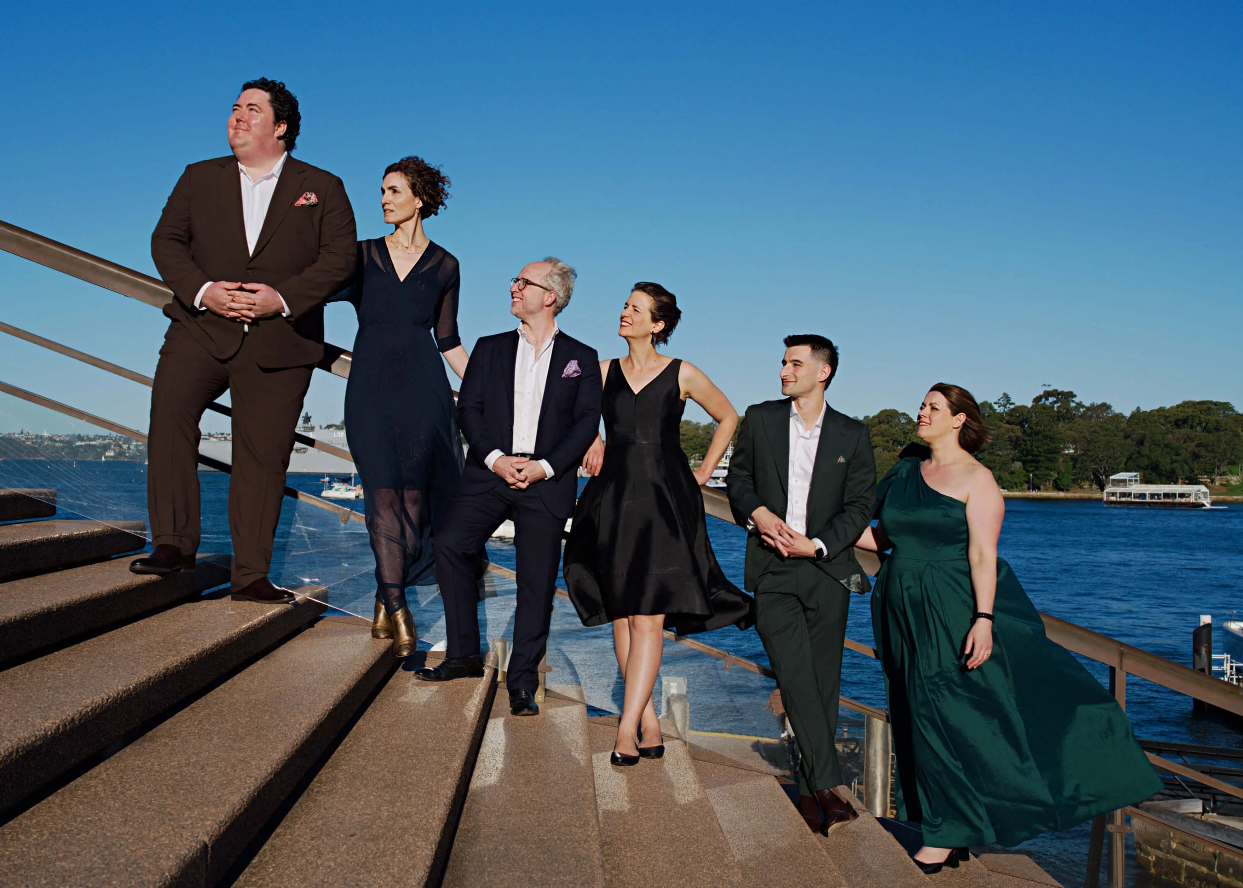 Group of six well-dressed people standing on outdoor steps near water with boats, harbor, and trees in the background on a clear day.