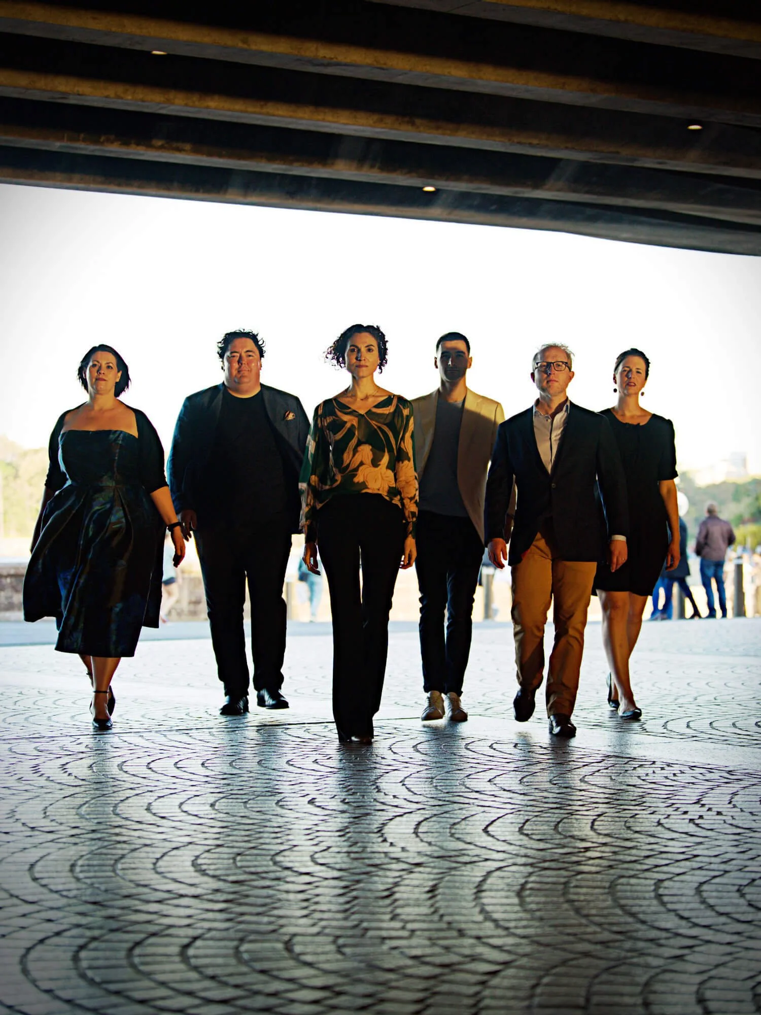 A group of six people walking outdoors under a bridge, with a patterned pavement and a clear sky in the background.