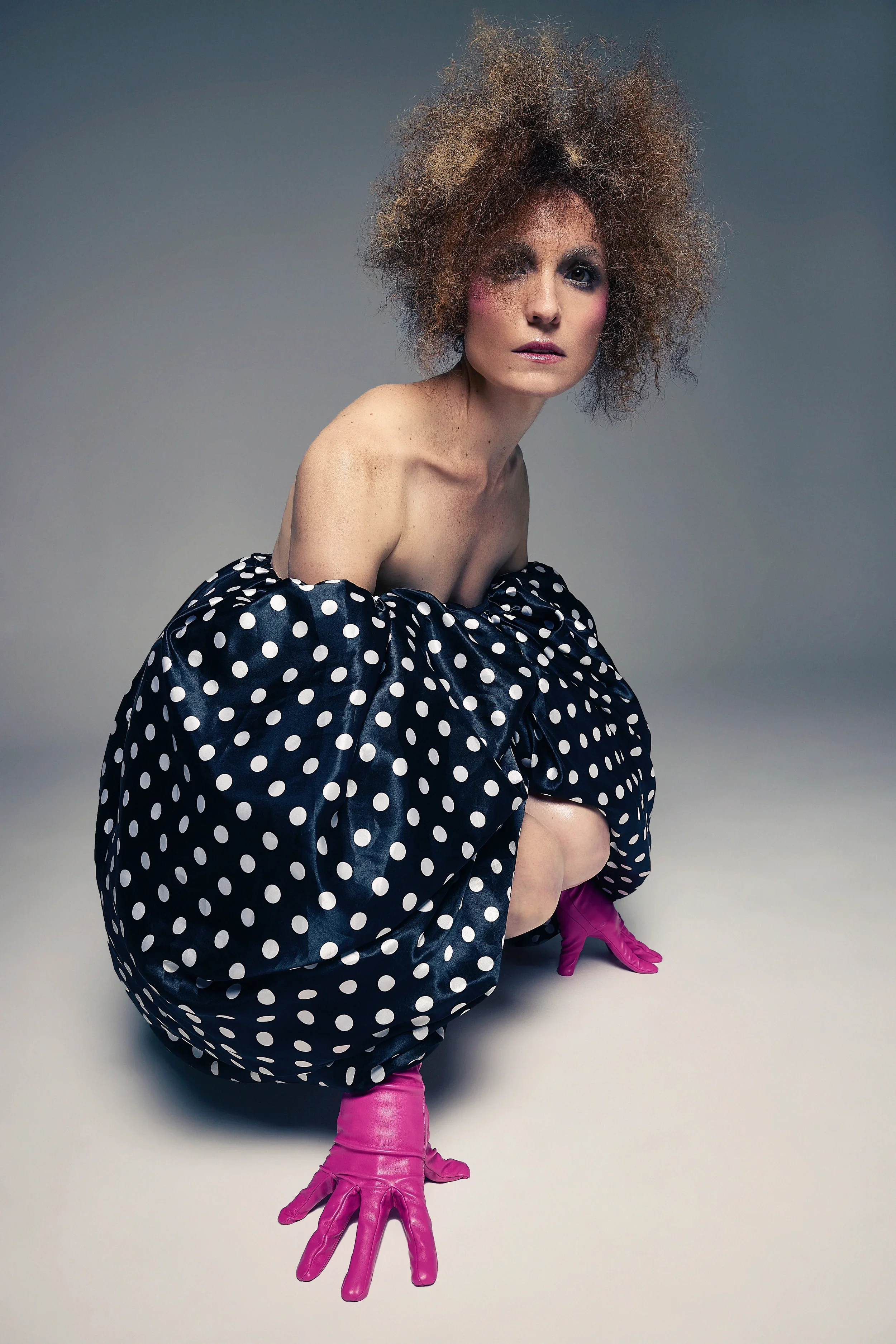 Jessica O'Donoghue with wild, curly hair and pink gloves is posed on the floor, wearing a polka dot dress, against a plain gray background.