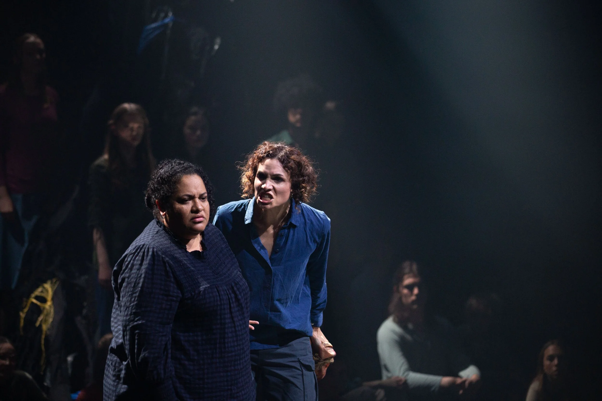 Two women on stage, one with curly black hair and dark skin, the other with curly brown hair and light skin, appear upset or angry. They are part of a group of people standing in the background, watching intensely.
