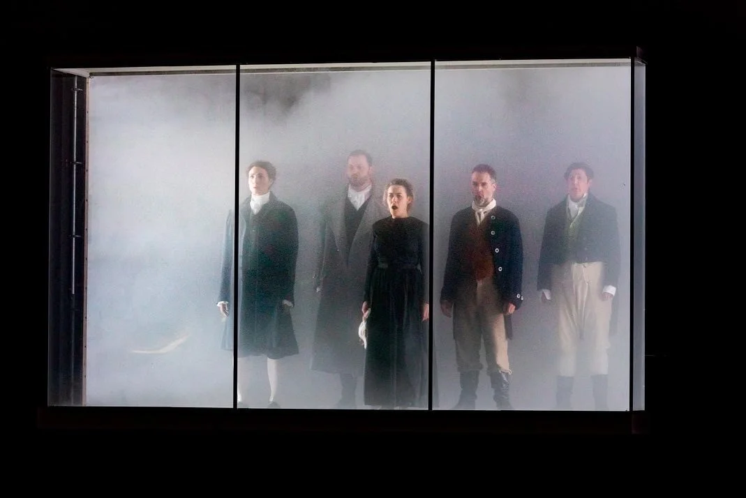 Five people dressed in historical clothing standing inside a glass display case filled with fog or mist, with a black background.