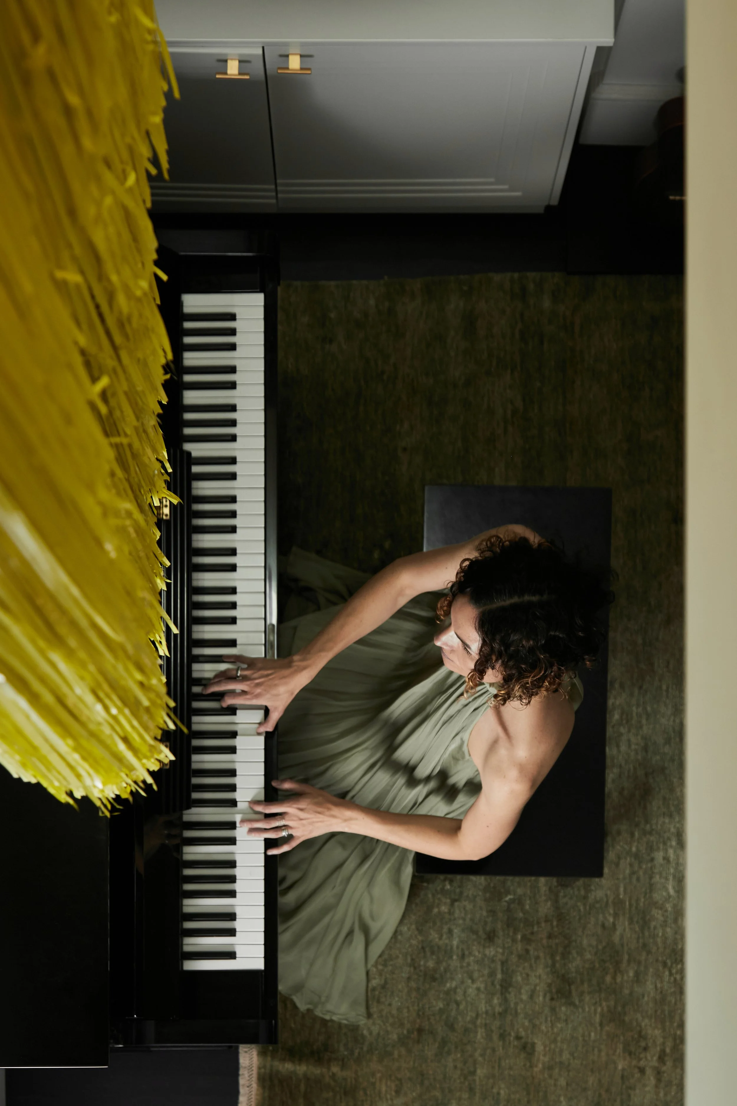 Jessica O'Donoghue with curly hair wearing a light green dress playing a black upright piano decorated with yellow paper flowers.