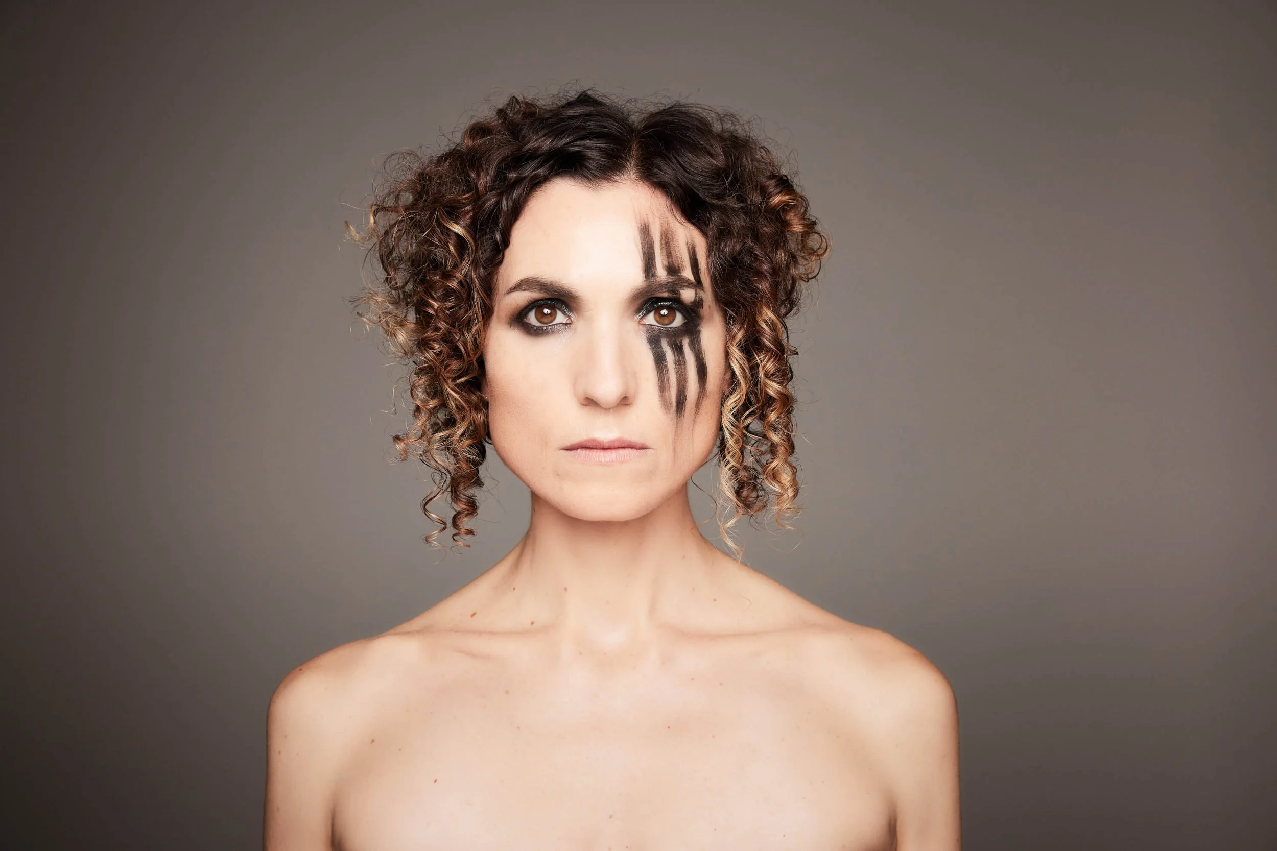 Jessica O'Donoghue with curly brown hair and brown eyes, with black painted lines on her face, stands against a gray background, with her shoulders bare.