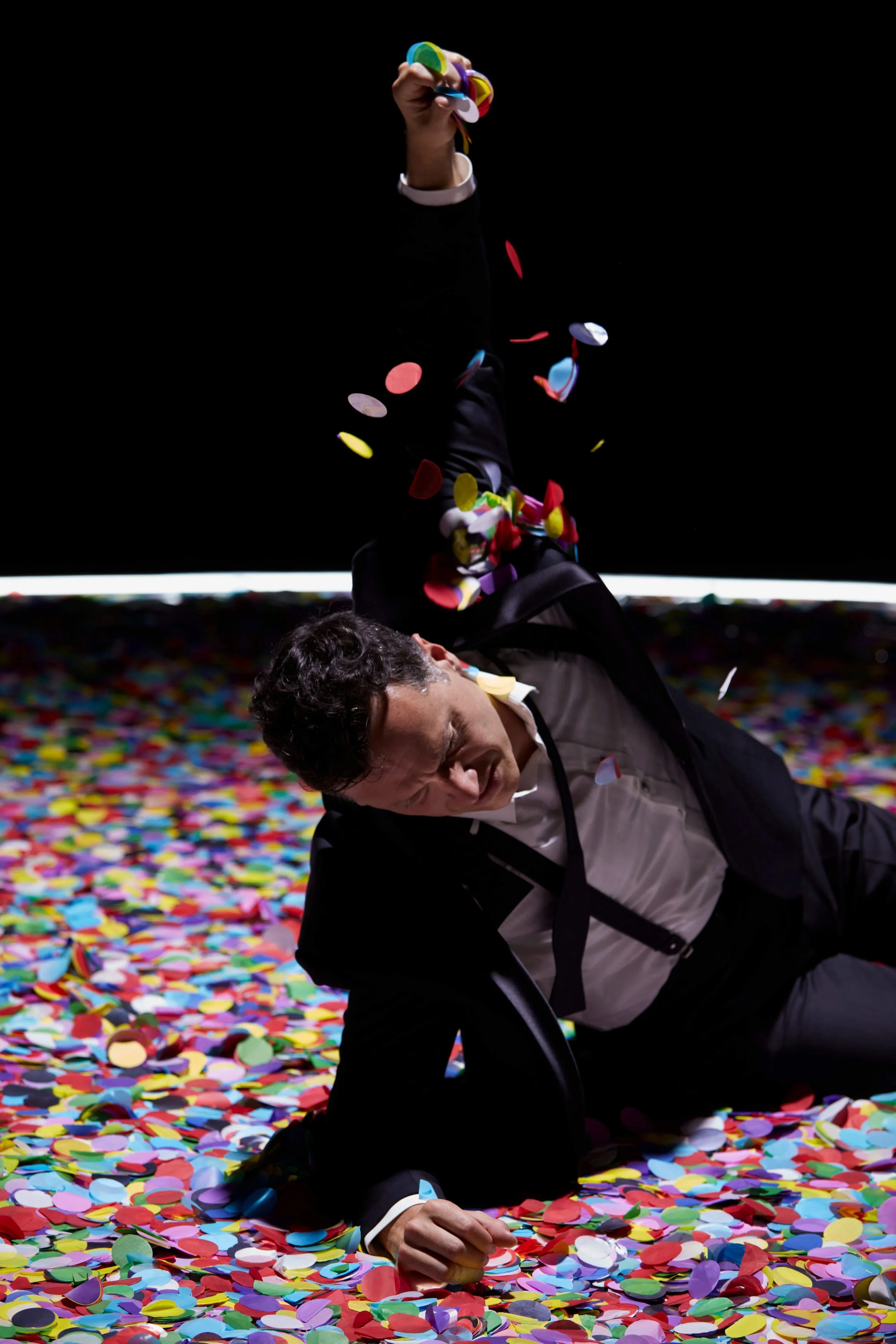 Two men in formal suits lie on a colorful confetti-covered floor, one on his side and one propped on his elbow, with multicolored confetti raining down around them in a dark setting.