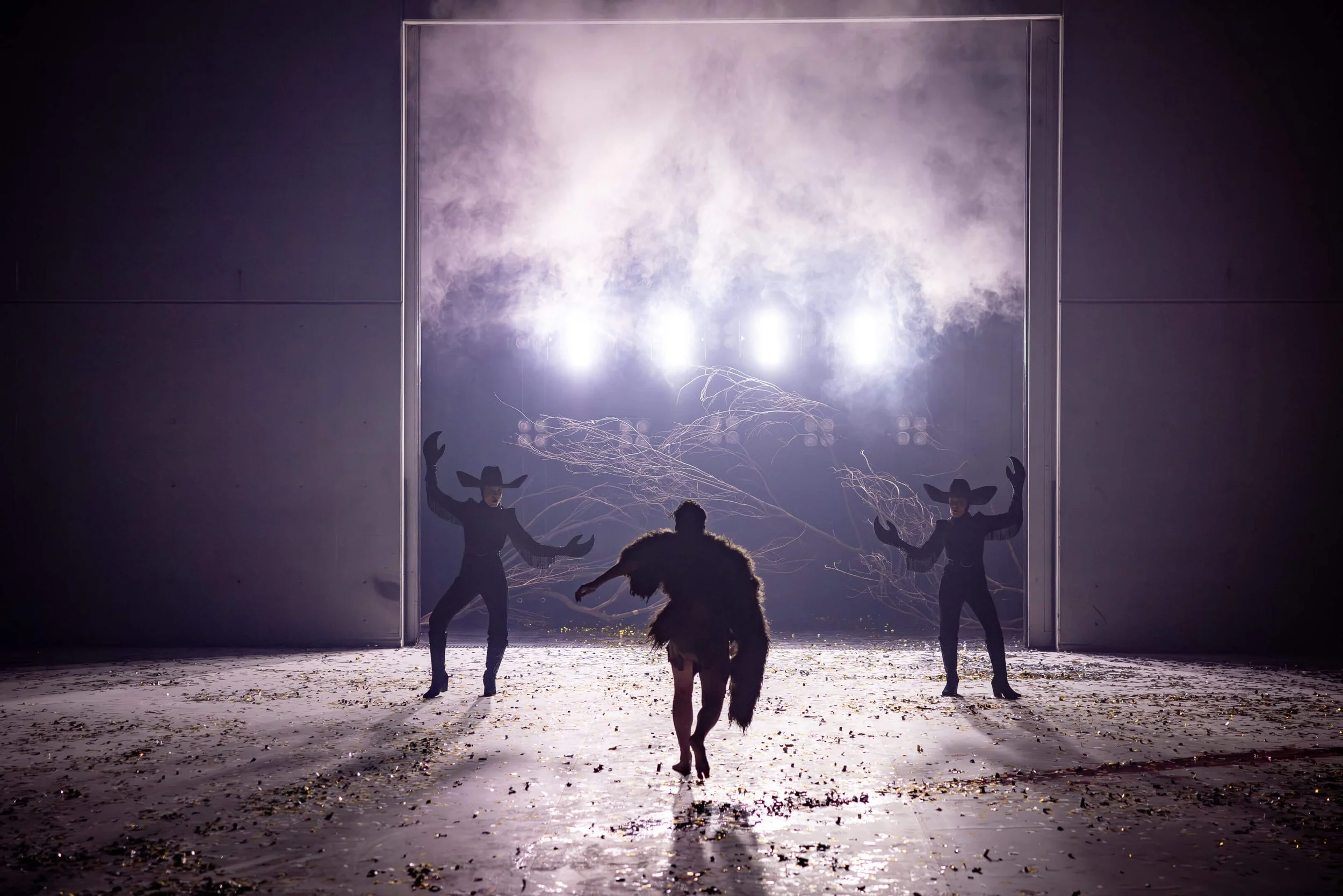 Performance scene with performers dressed in black costumes with large headpieces, performing in front of a glowing backdrop with lightning effects, on stage with an audience