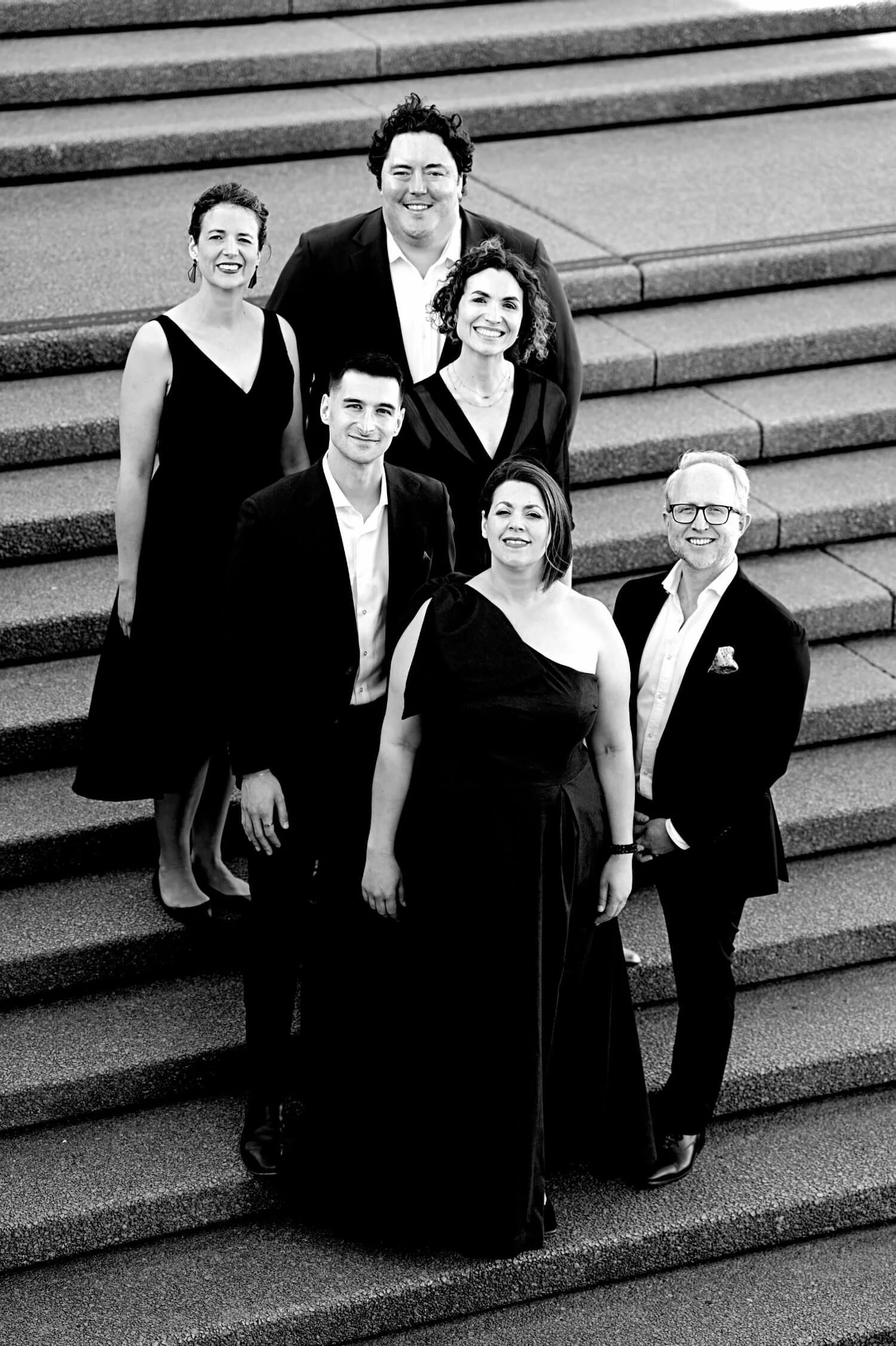 A group of seven people dressed in formal attire standing on steps, smiling for a group photo, with four women and three men, in black and white.