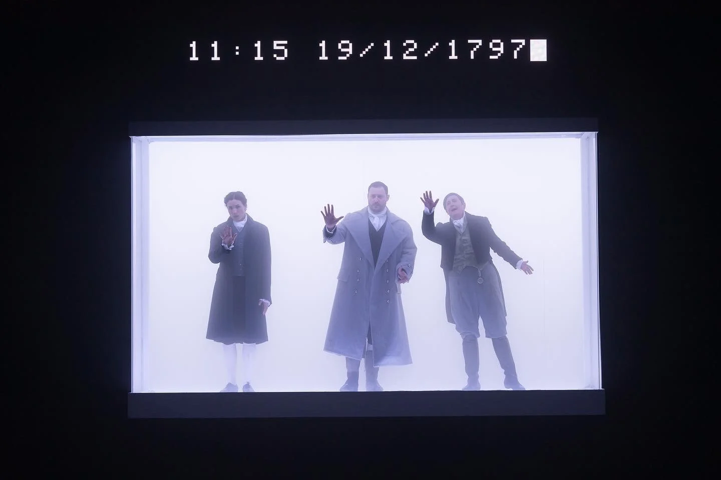 Three men and one woman in vintage clothing standing behind a glass case, illuminated by bright white light, with their hands pressed against the glass, as if trapped inside.