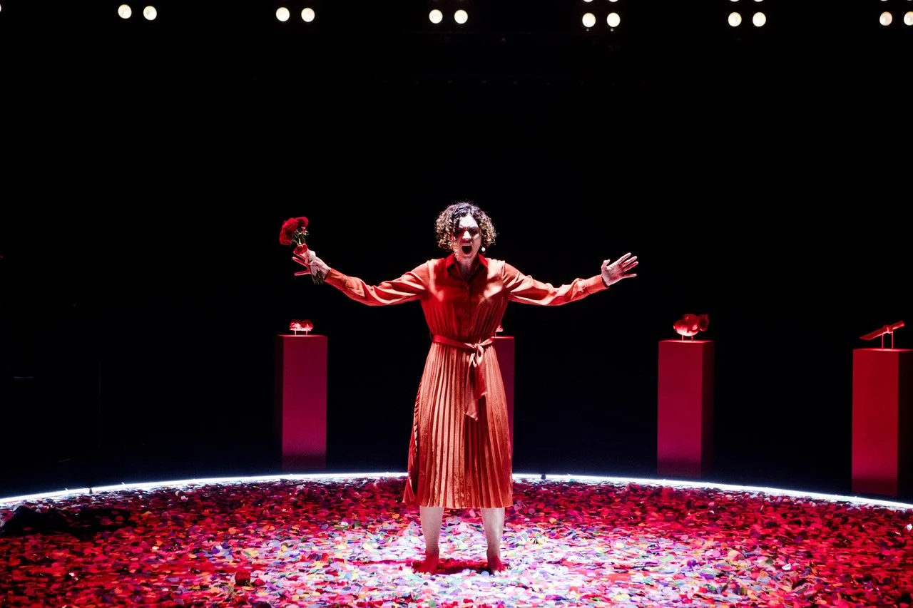 Jessica O'Donoghue in a red dress stands on a stage covered in confetti, facing the audience with arms outstretched, holding a red rose in her left hand. 