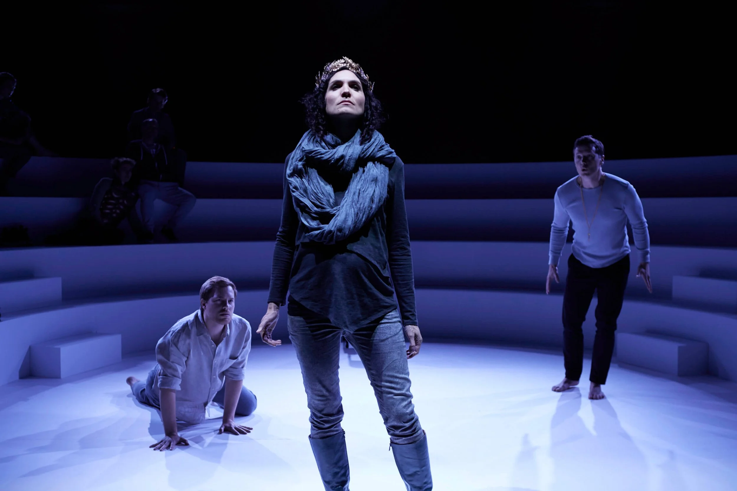 Group of people performing a theatrical scene on stage, with Jessica O'Donoghue wearing a crown and scarf, standing in the foreground; others are kneeling or standing behind her in a dark setting.