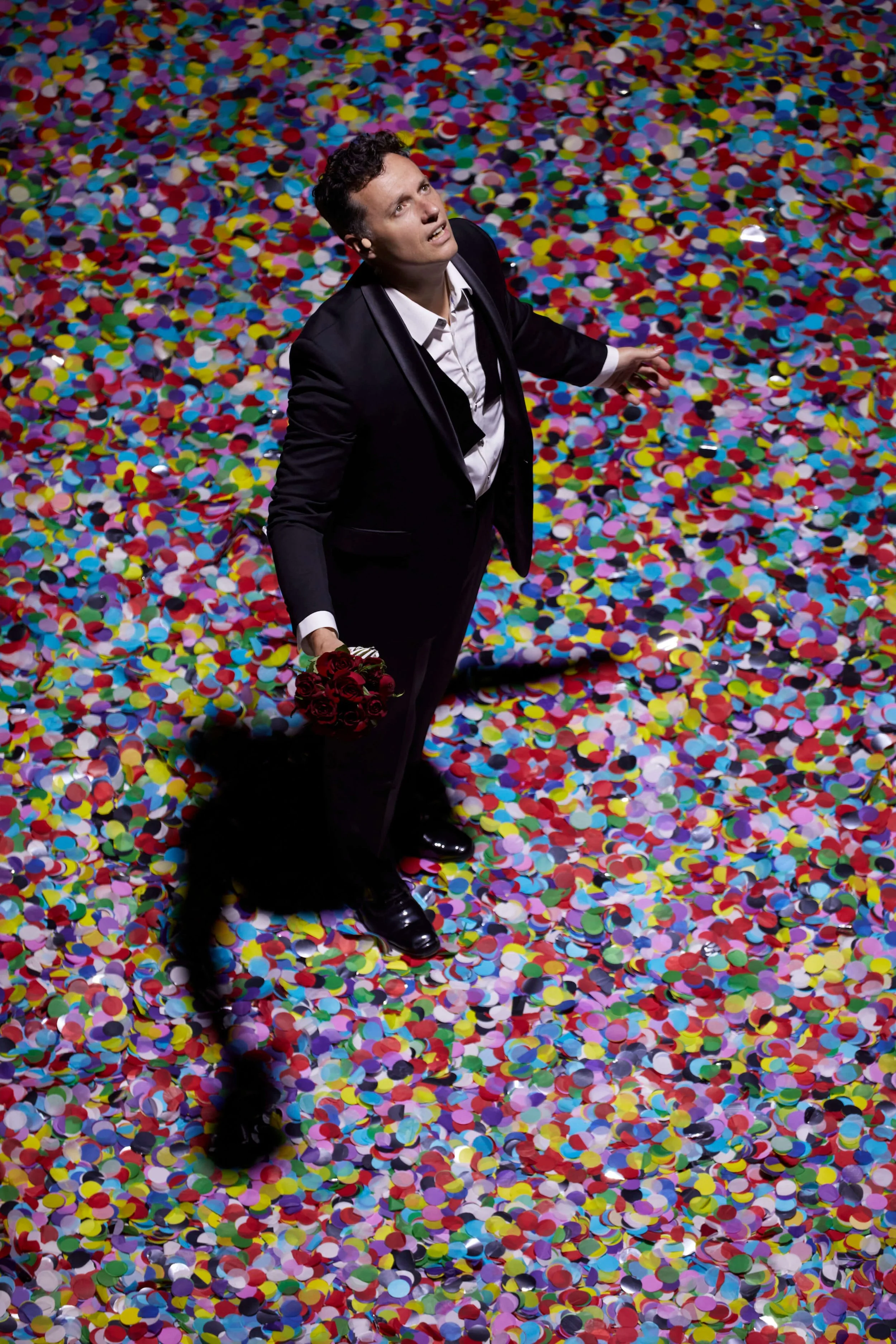 A man in a black tuxedo holding a bouquet of red roses on a colorful confetti-covered floor, looking up with a dramatic expression.