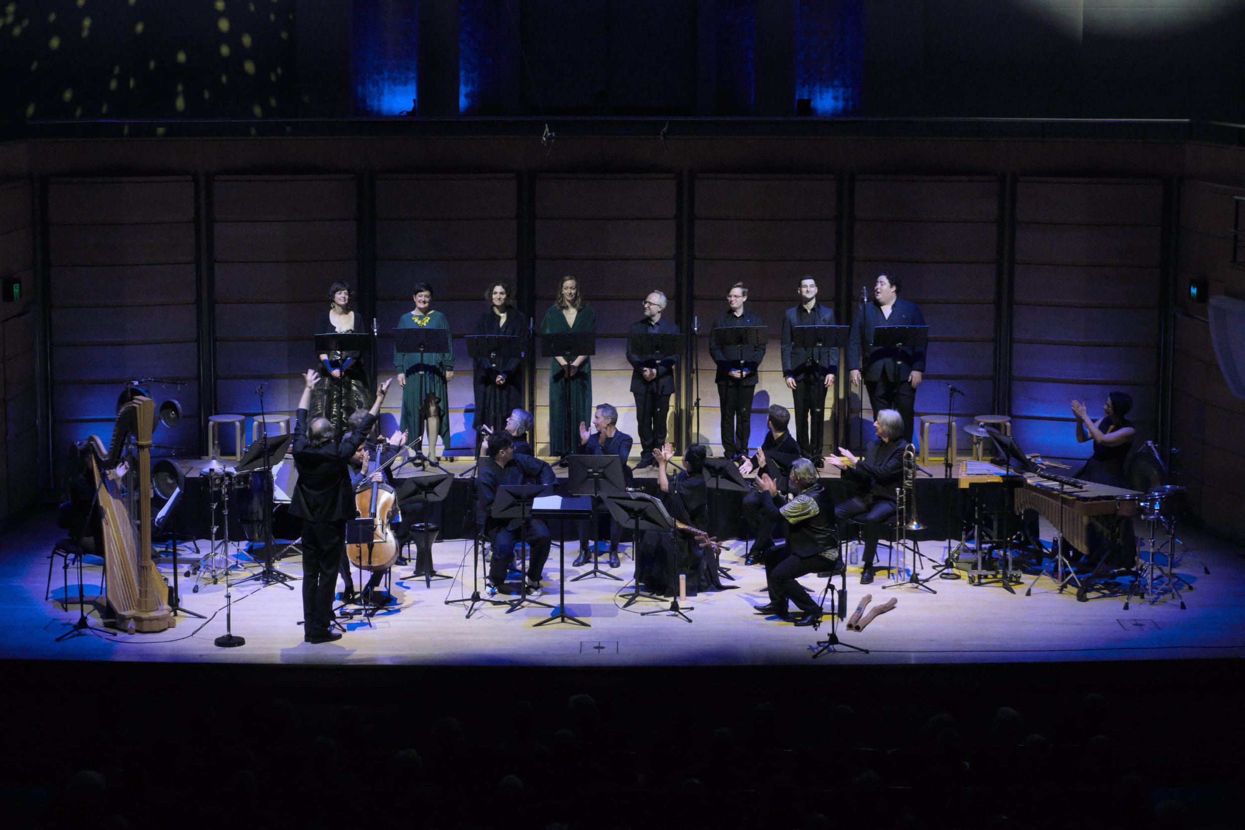 Orchestra and choir performing on stage in a concert hall, conducted by a conductor, with various musical instruments visible.