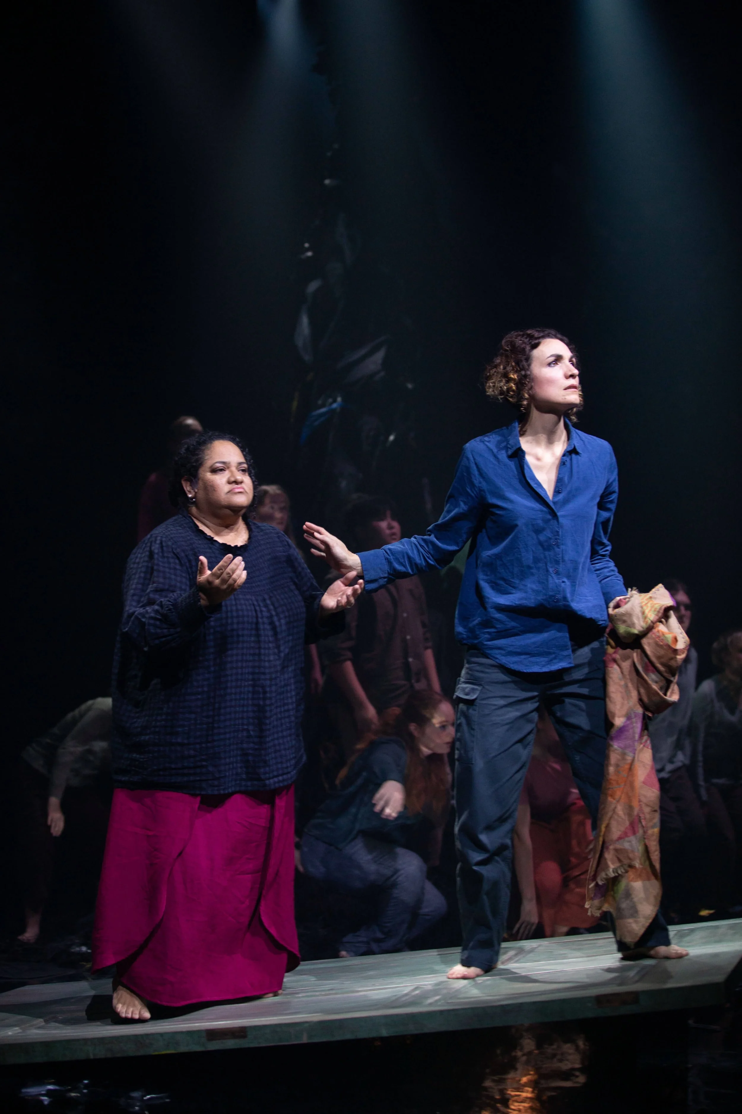 Two women standing on a stage with dark background, surrounded by other people, one in bright pink skirts and the other in a blue shirt and jeans, appearing to perform or participate in a theatrical or artistic event.