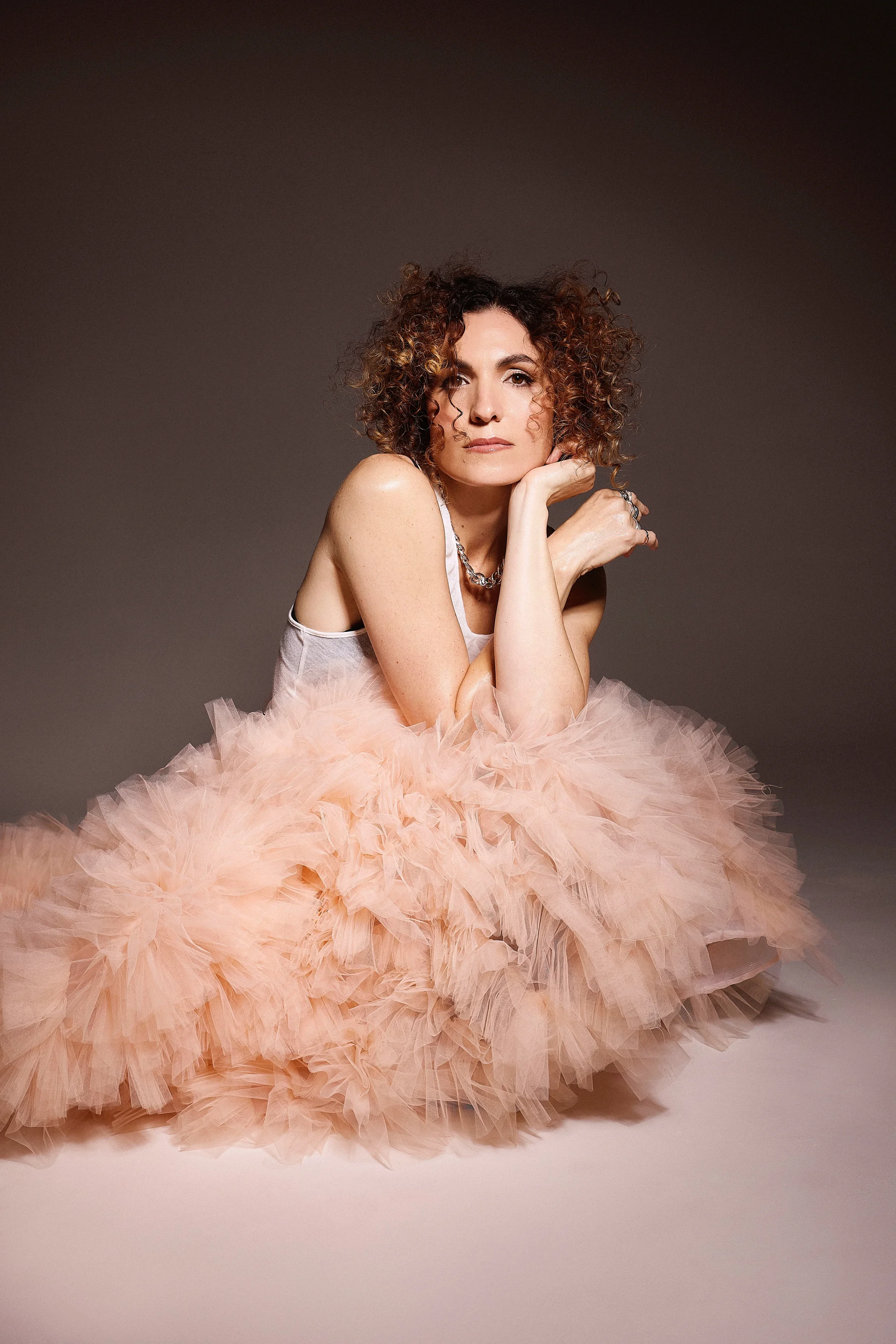 Jessica O'Donoghue with curly hair and light skin wearing a white top and a large, fluffy pink tulle skirt, sitting with her chin resting on her hand, looking at the camera against a plain background.
