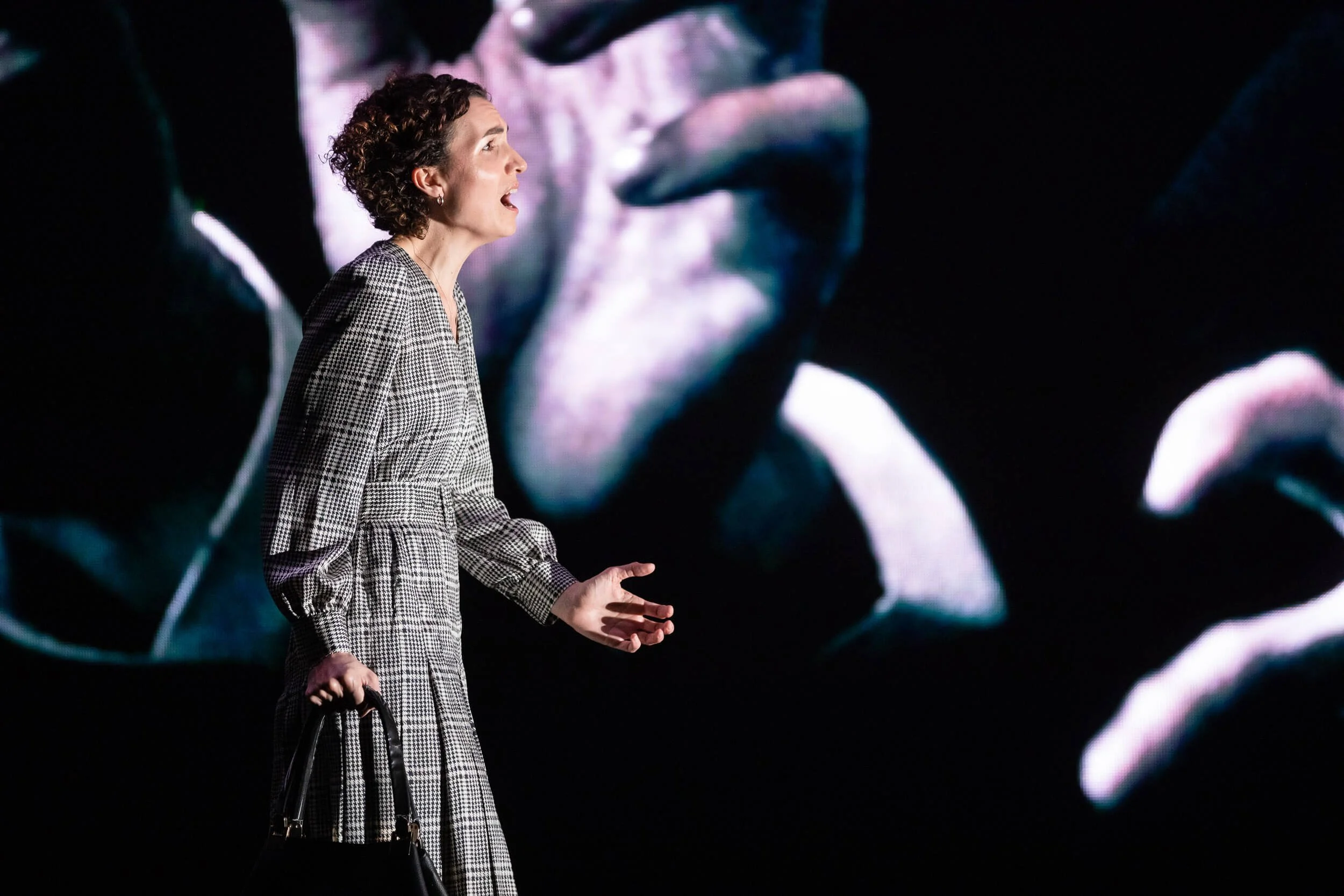 Jessica O'Donoghue with short, curly hair wearing a checkered dress is speaking on stage with her hands raised. Behind her is a large screen displaying a close-up image of her face.
