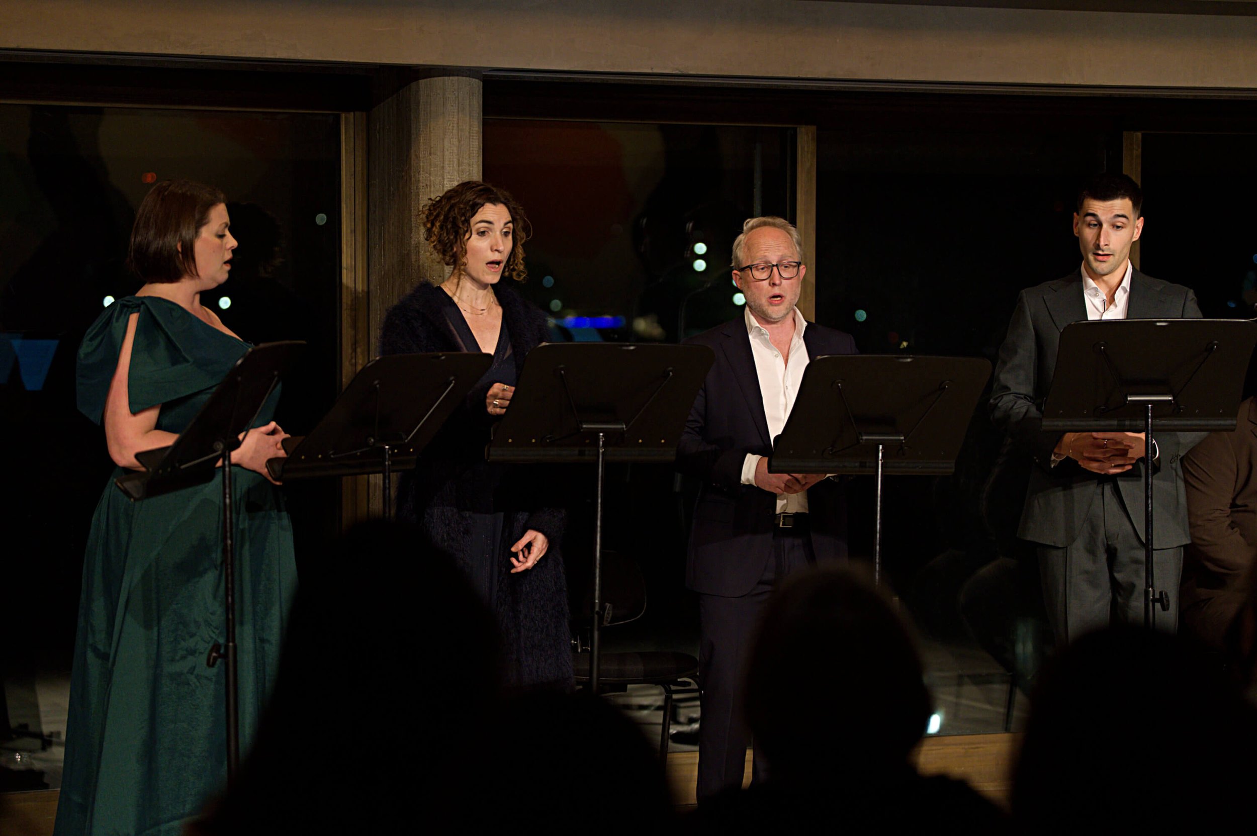 Four people standing in front of music stands, singing or performing in a room with large windows at night.