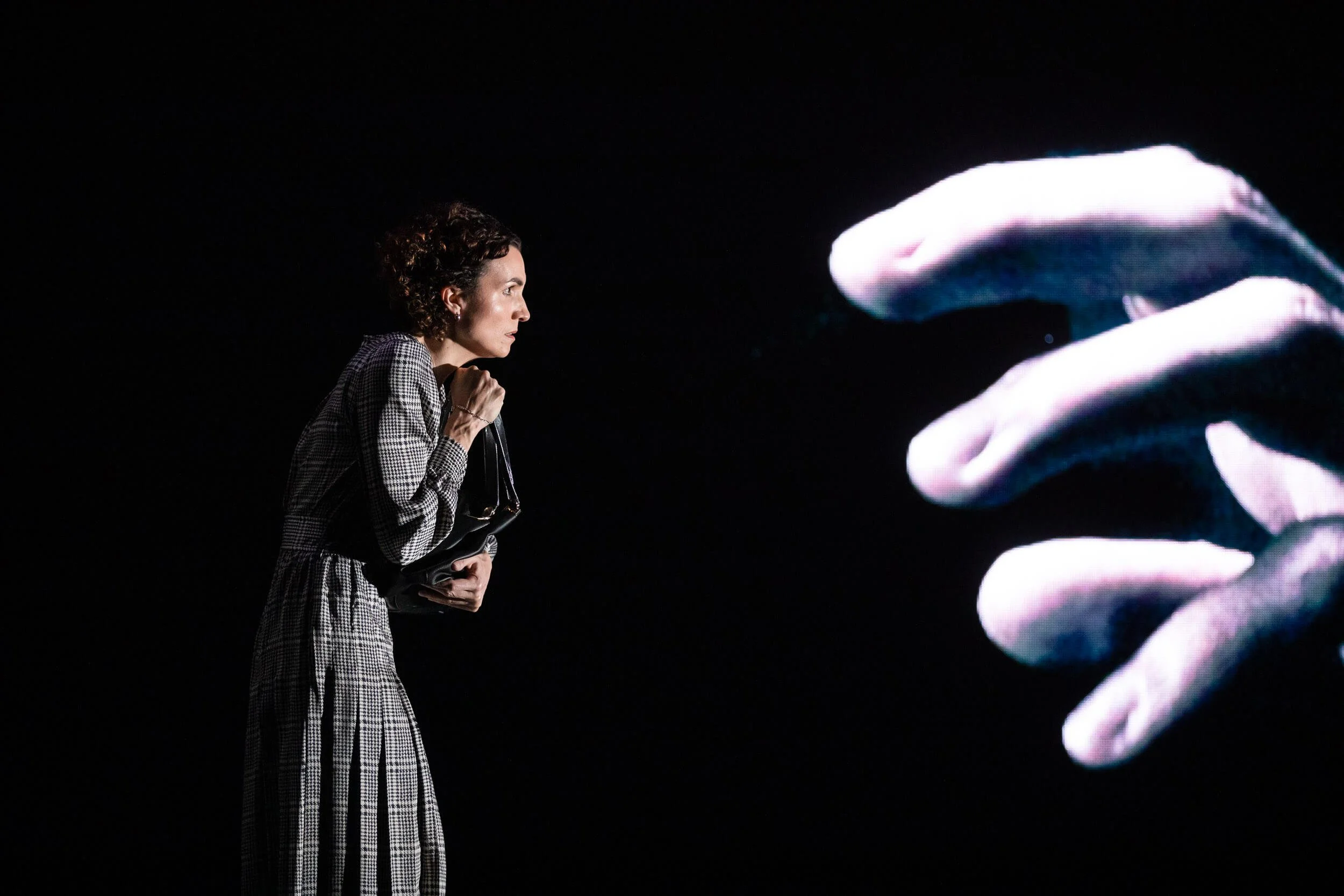 Jessica O'Donoghue with short curly hair wearing a checkered outfit, holding a purse, looking at a large digital display with abstract hand-like shapes.
