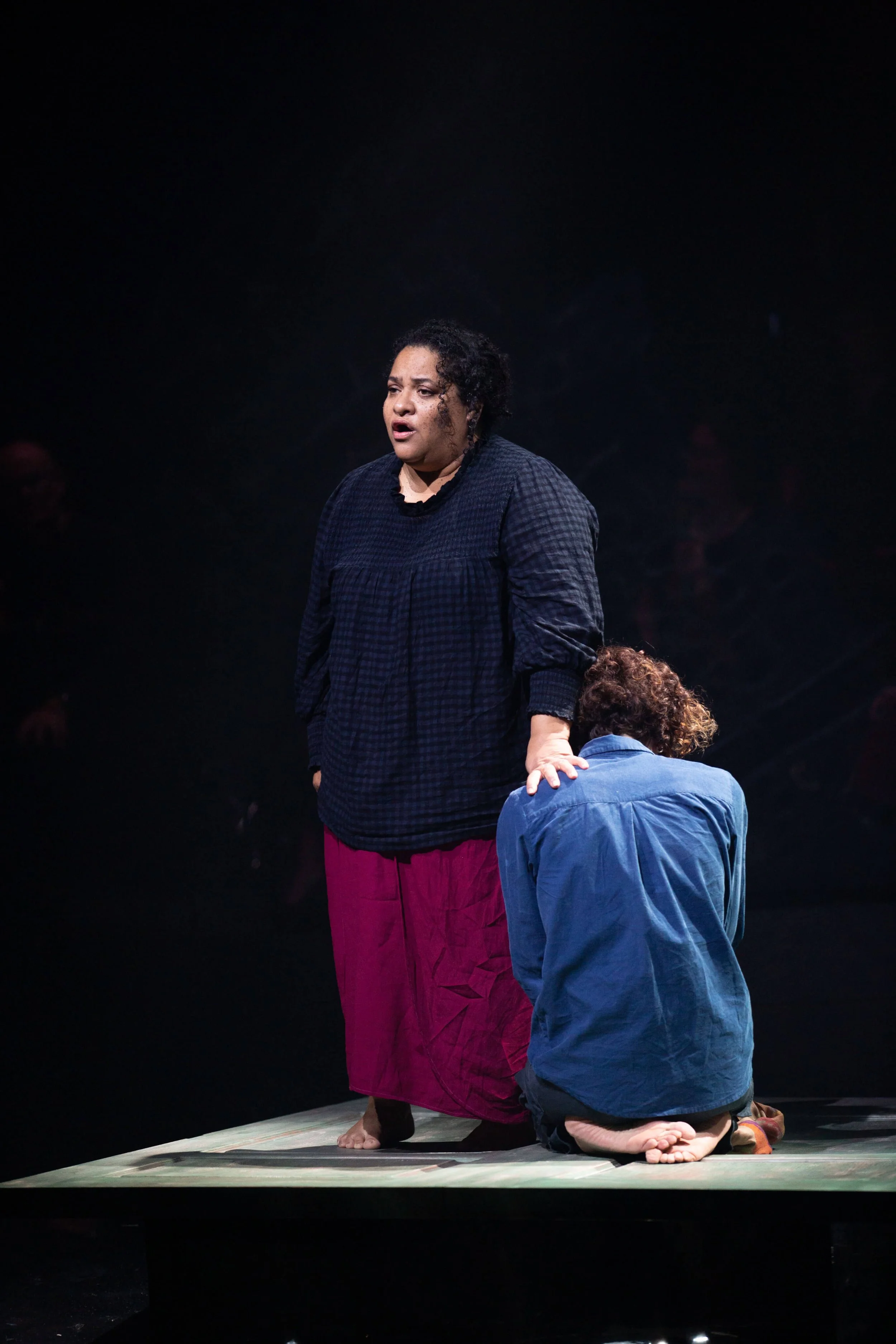 A woman with curly dark hair standing on stage, appearing emotional, next to a person kneeling with back facing the camera.