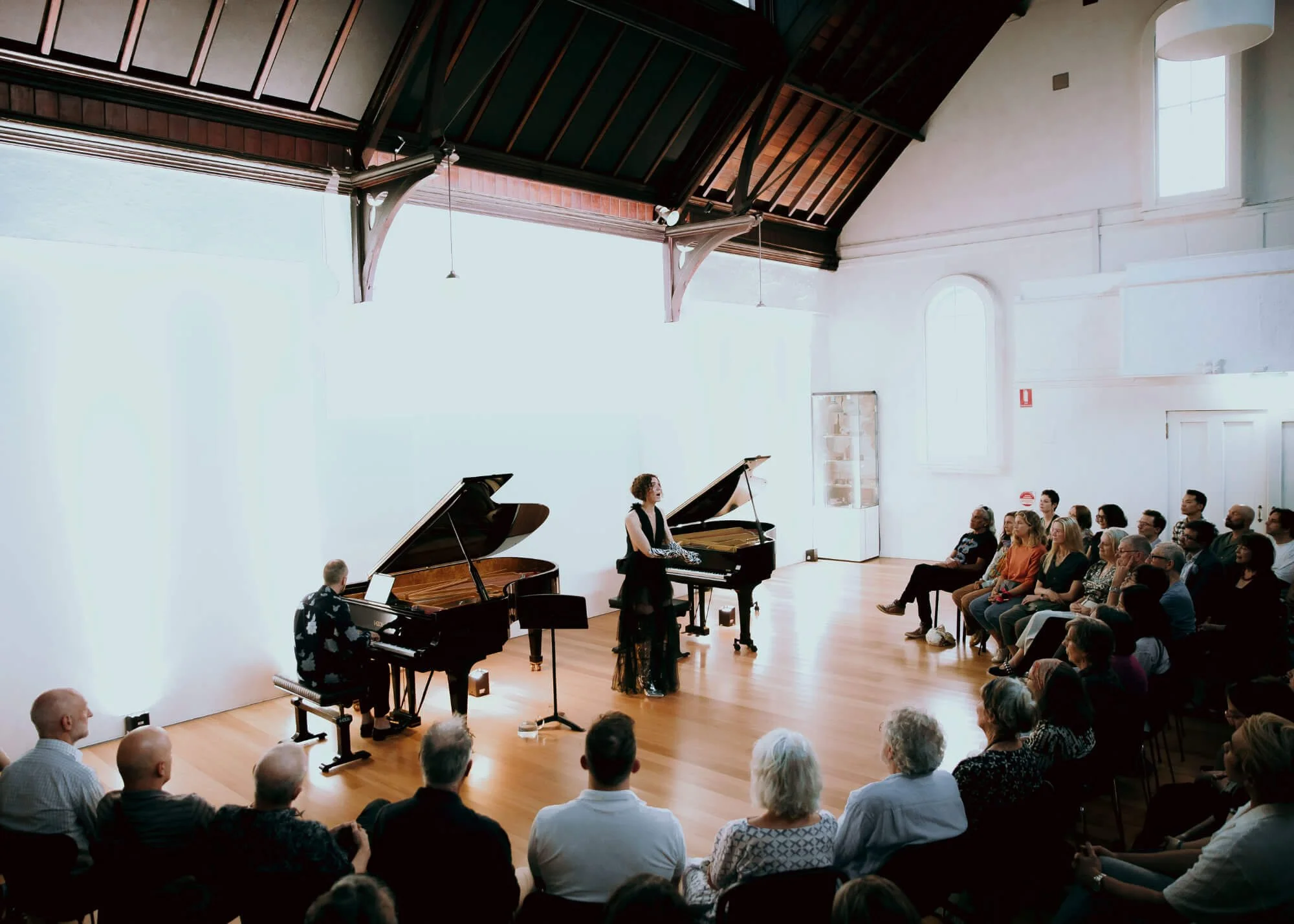 Jessica O'Donoghue performing on a grand piano while a man plays another grand piano in a spacious room with a high ceiling and wooden beams, with an audience sitting and watching.