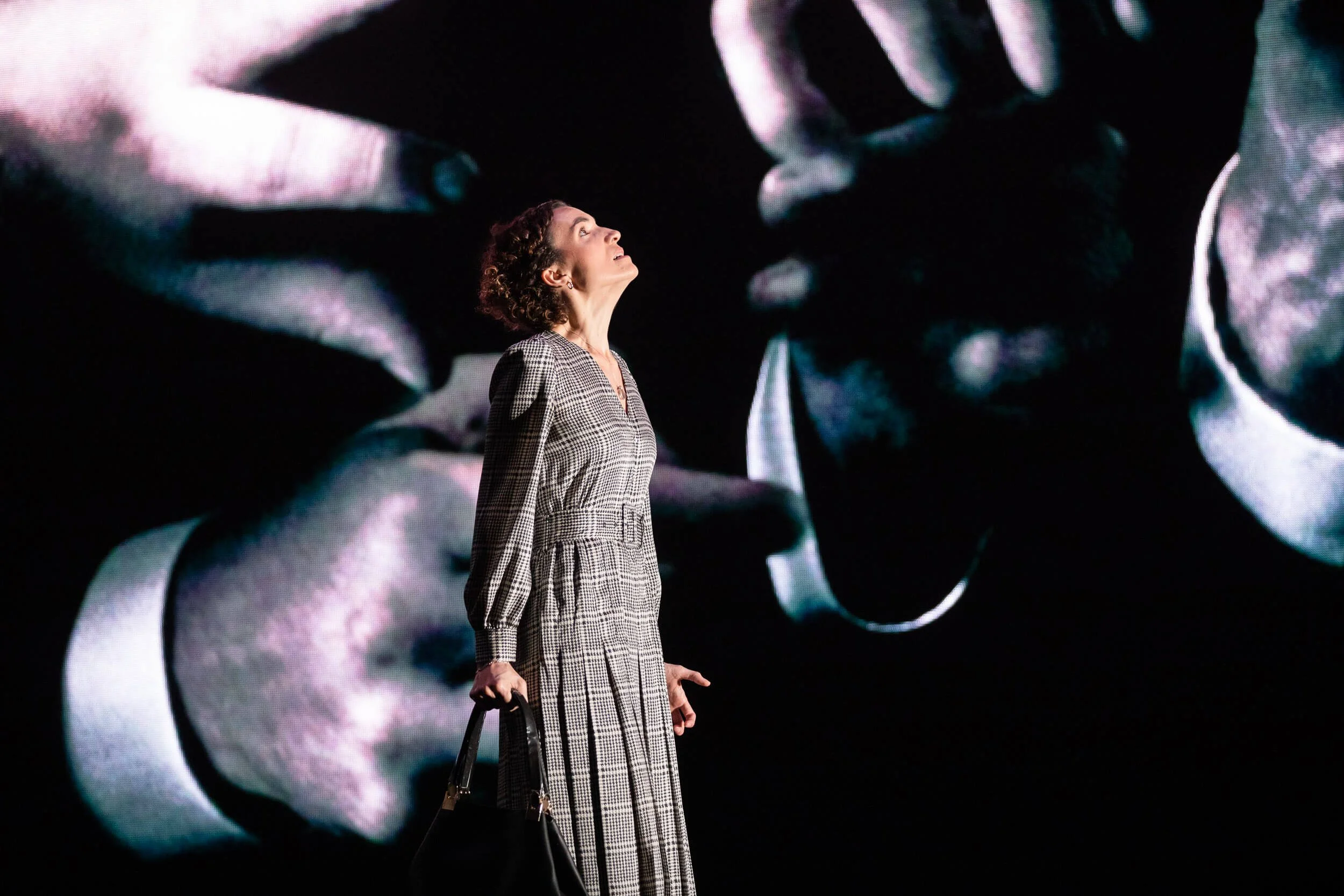 Jessica O'Donoghue in plaid dress standing in front of a large screen displaying abstract shapes, holding a handbag, looking upward.