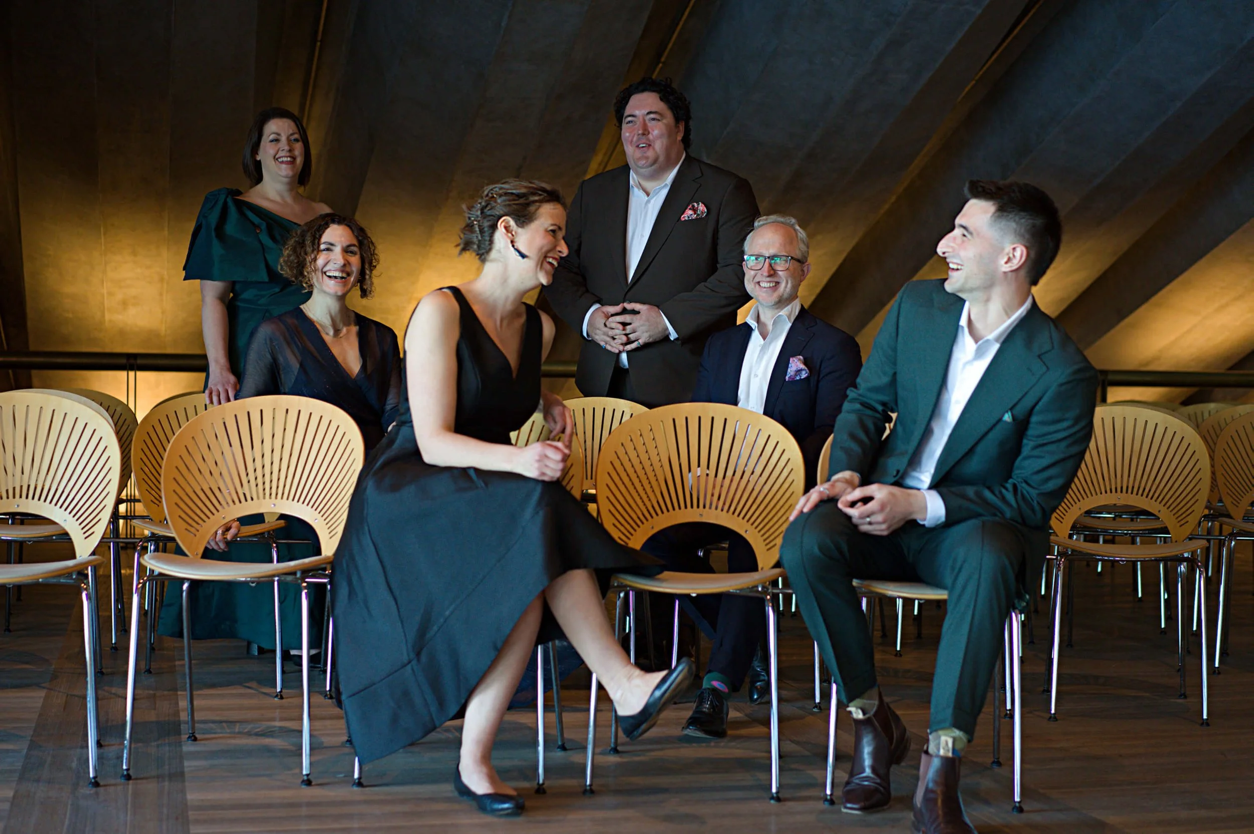 Group of six people in formal attire sitting and standing in a modern, warmly lit event space with wooden chairs, engaging in conversation and laughing.