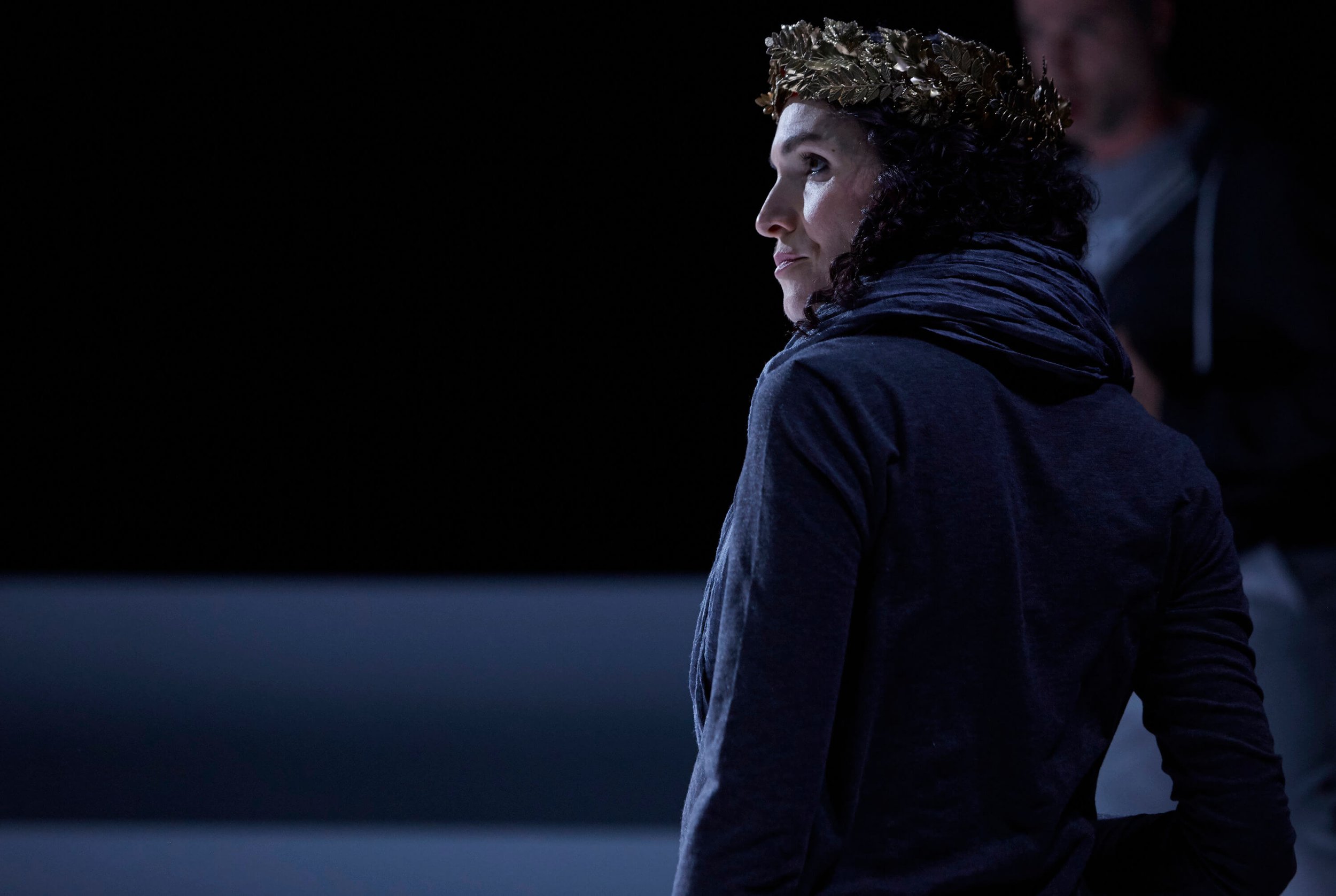 Jessica O'Donoghue with curly hair wearing a gold laurel wreath on her head, looking to the side, dressed in a dark jacket, in a dark environment.