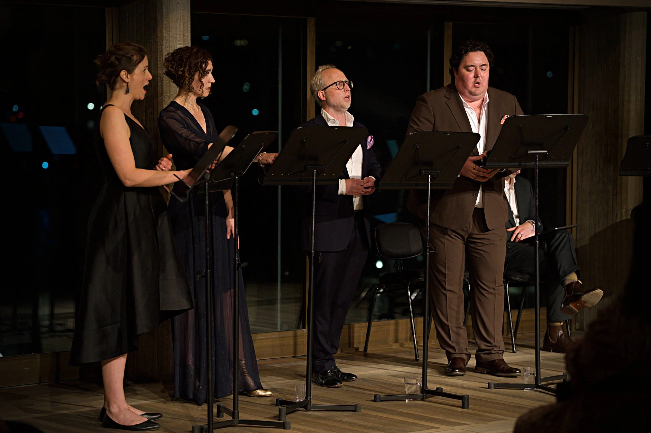 Group of five actors performing in a theater, standing behind music stands, with their mouths open as if singing or speaking.