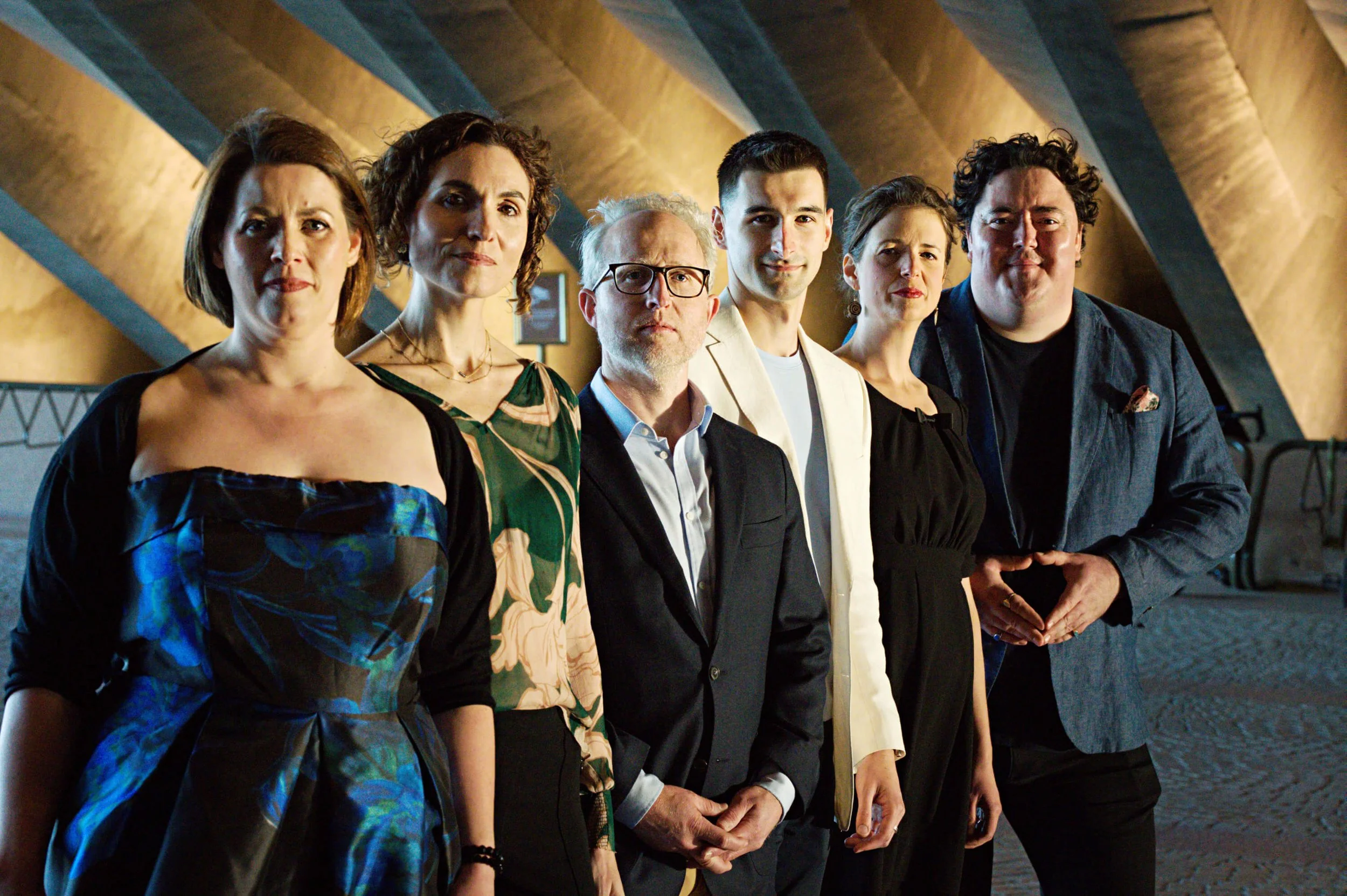 Group of seven diverse adults standing together indoors against a textured wooden wall, dressed in business casual attire, looking at the camera with neutral or slight smiles.