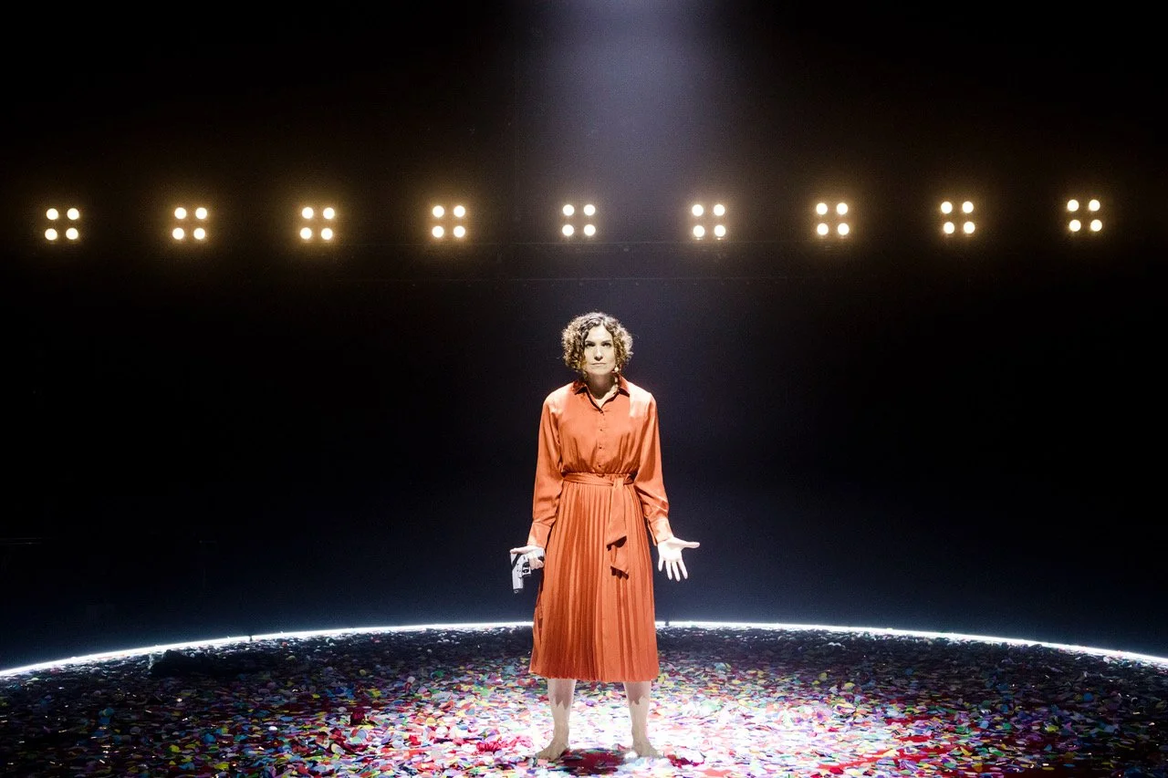 Jessica O'Donoghue stands on a stage with scattered colorful confetti, wearing an orange dress, under bright stage lights in a dark background.