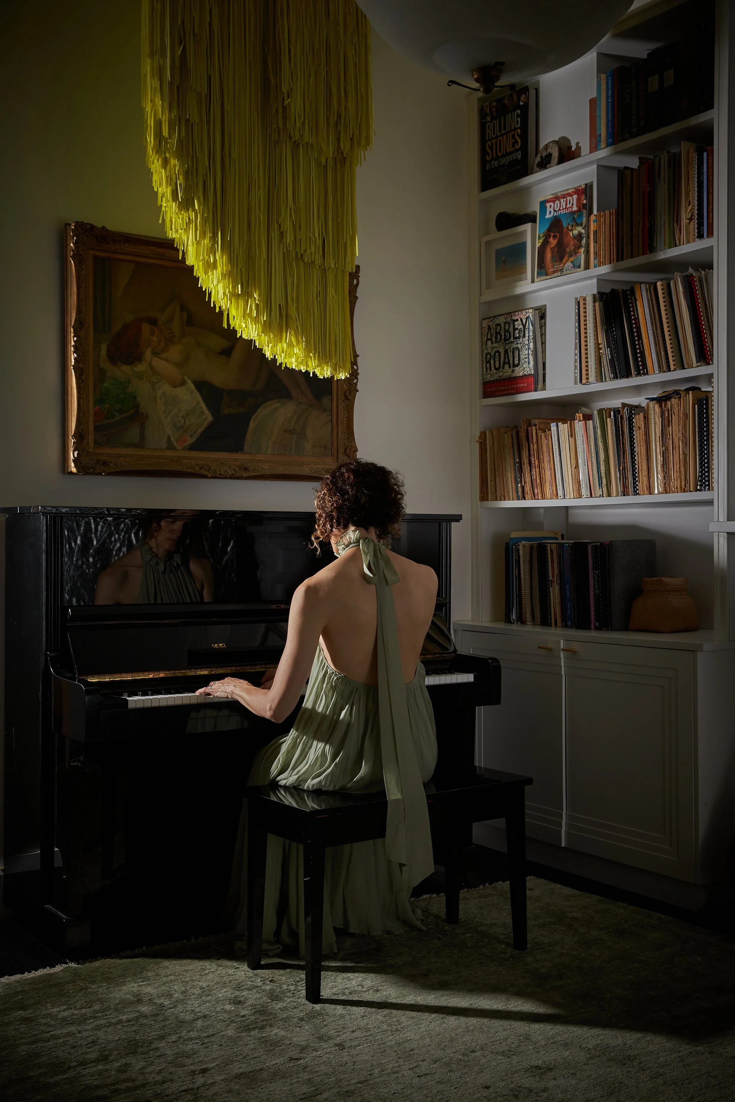 A woman in a flowing, light-colored dress with a large bow at the back, playing a black grand piano in a dimly lit room. The room features white bookshelves with various books and framed artwork, and a large painting on the wall, with a yellow fringe-like chandelier hanging overhead.