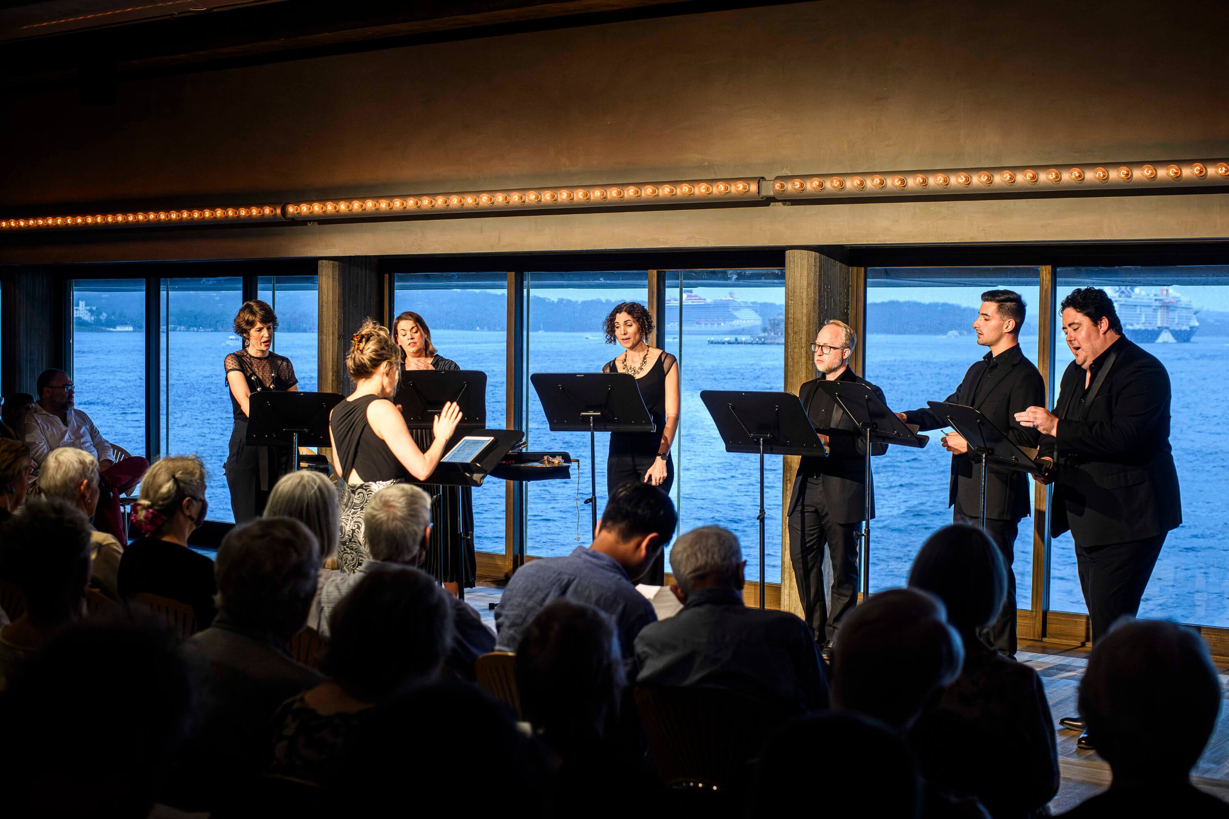 A group of singers performing on a stage with a large window showing water and ships in the background.