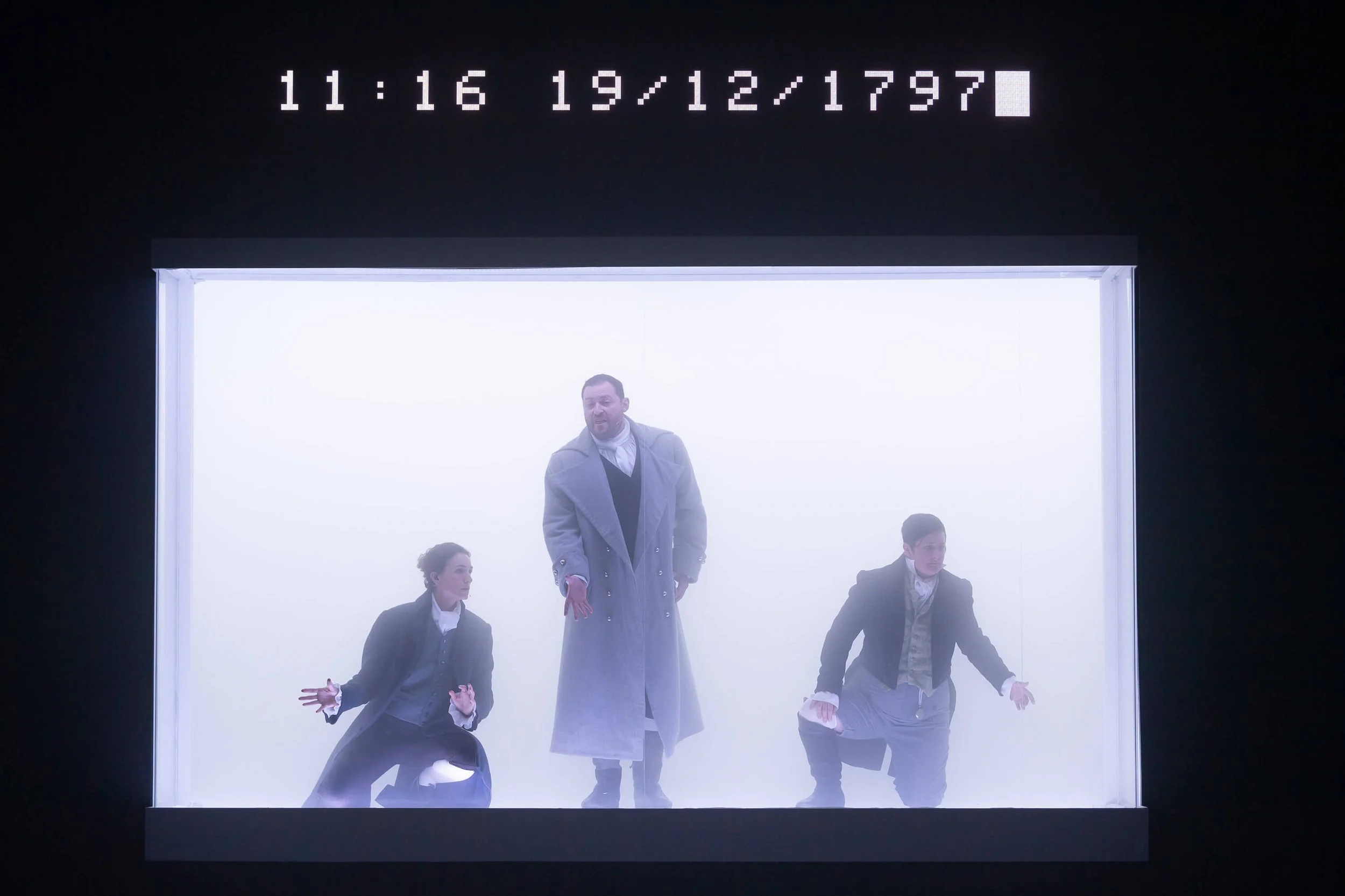 A group of four people, three men and one woman, are inside a rectangular, illuminated display box. The woman and two men are crouching, while another man is standing behind them. The background is misty or foggy, and there is a digital clock above d
