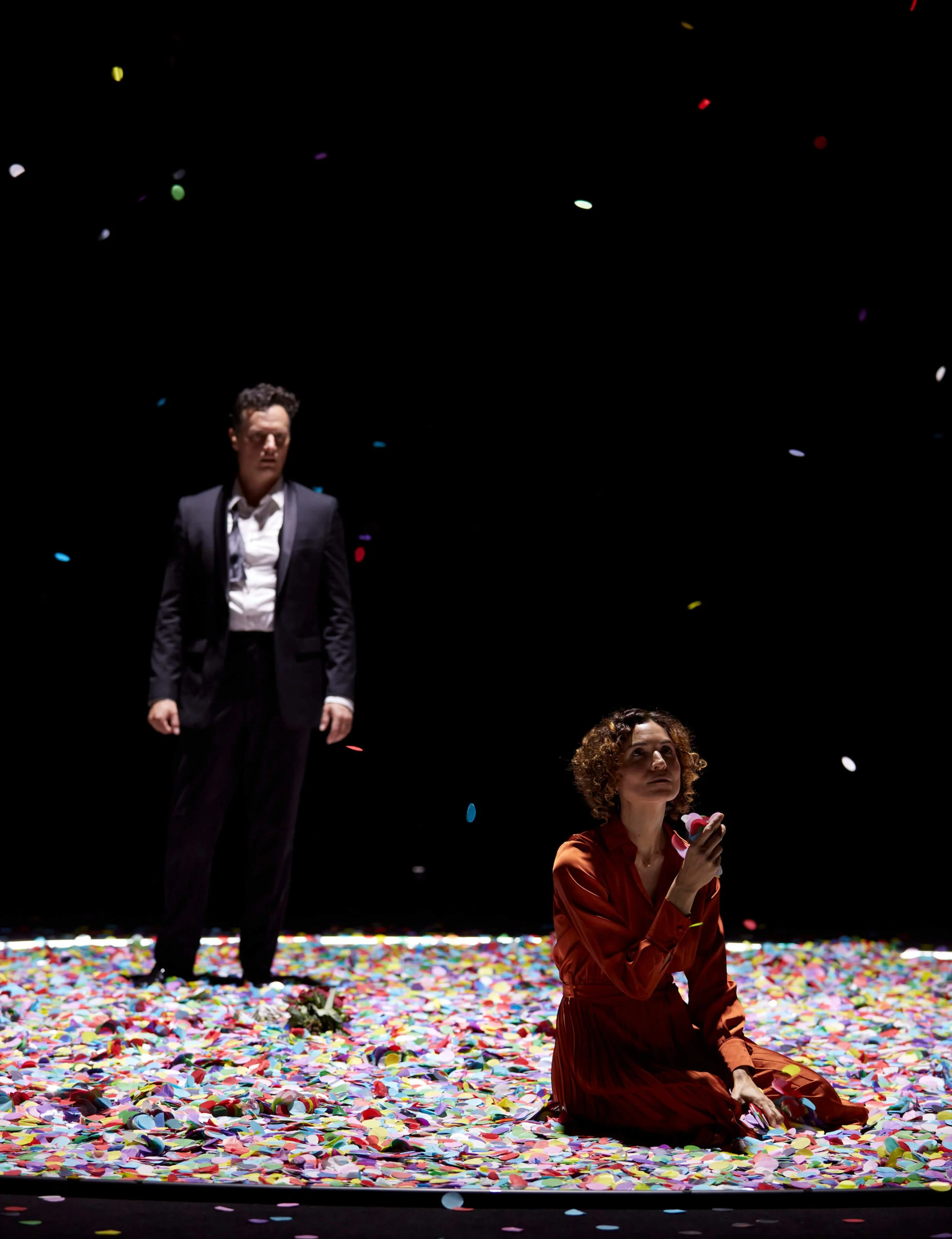 A man in a black suit and white shirt standing on a stage covered in colorful confetti, looking down with a serious expression, with Jessica O'Donoghue in a red dress sitting on the stage and holding confetti in her hand.