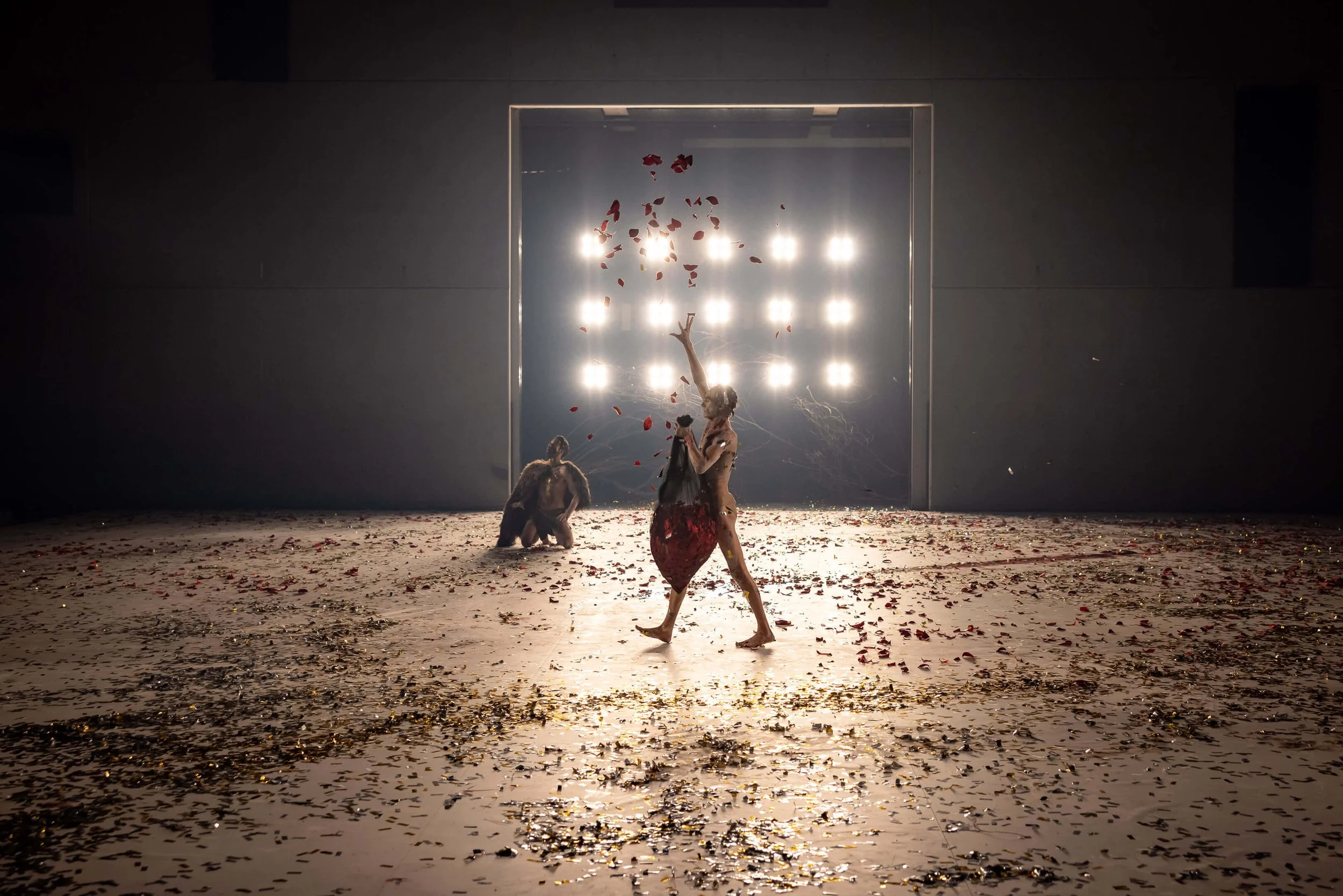 Two dancers perform on a stage covered with petals, backlit by bright lights, with a minimalistic gray background.