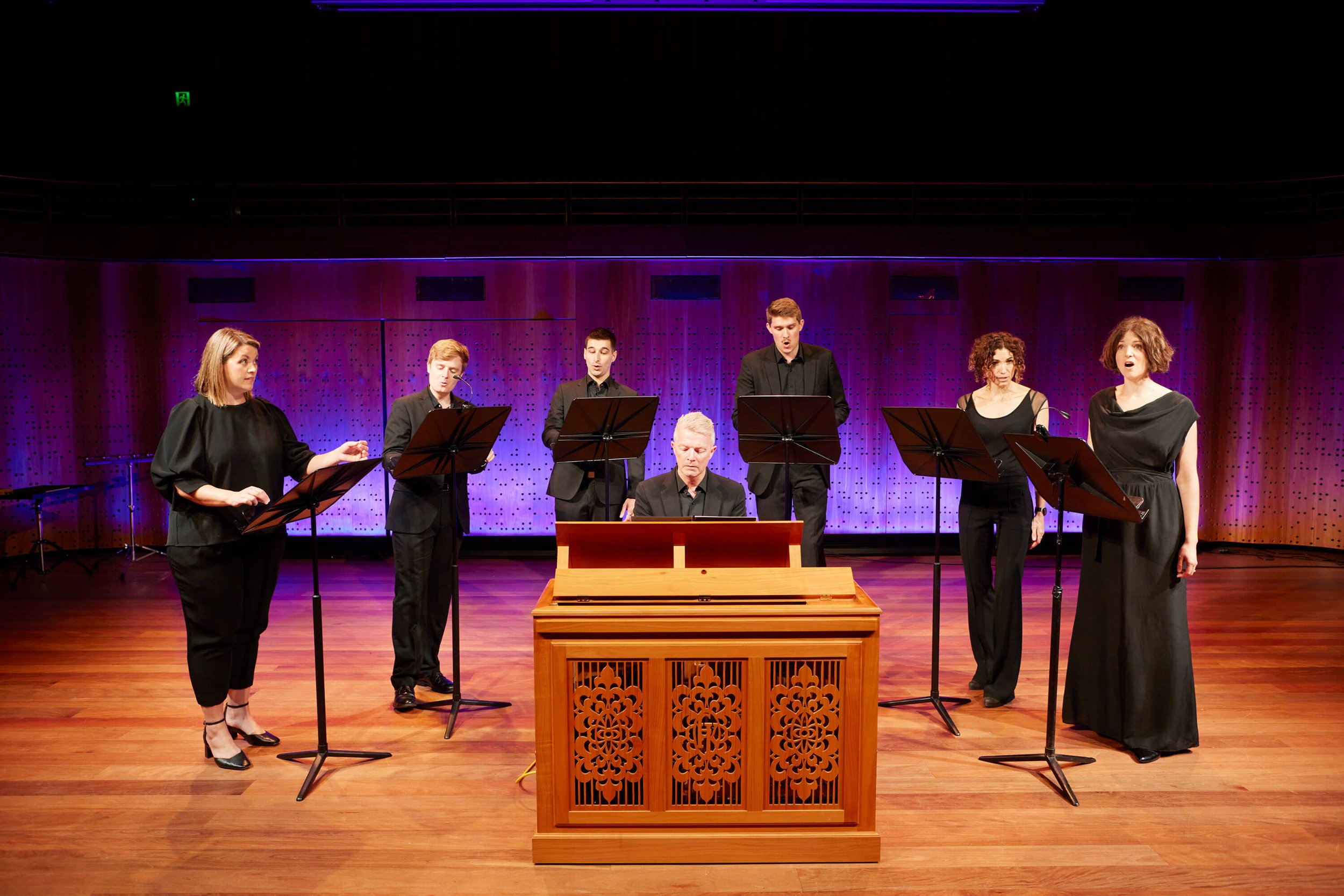 Group of six performers, three men and three women, singing on stage with music stands, a pianist playing a keyboard, and a wooden decorative music stand in front of them, against a purple-lit wooden backdrop.