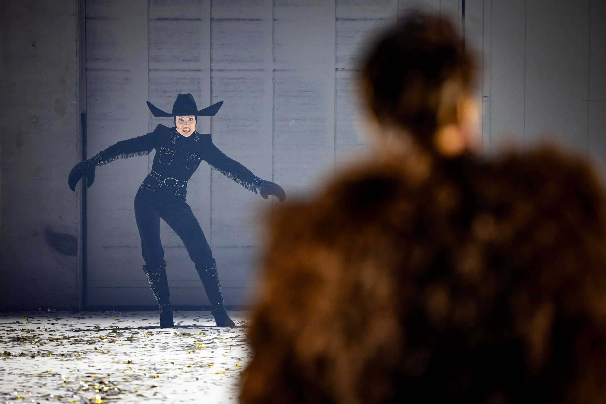 A woman dressed as a witch with a large black hat, black gloves, and a black fringed outfit appears frightened or angry as she faces an unfamiliar person. The scene is set against a gray wall with a floor scattered with yellow leaves, in a dimly lit 