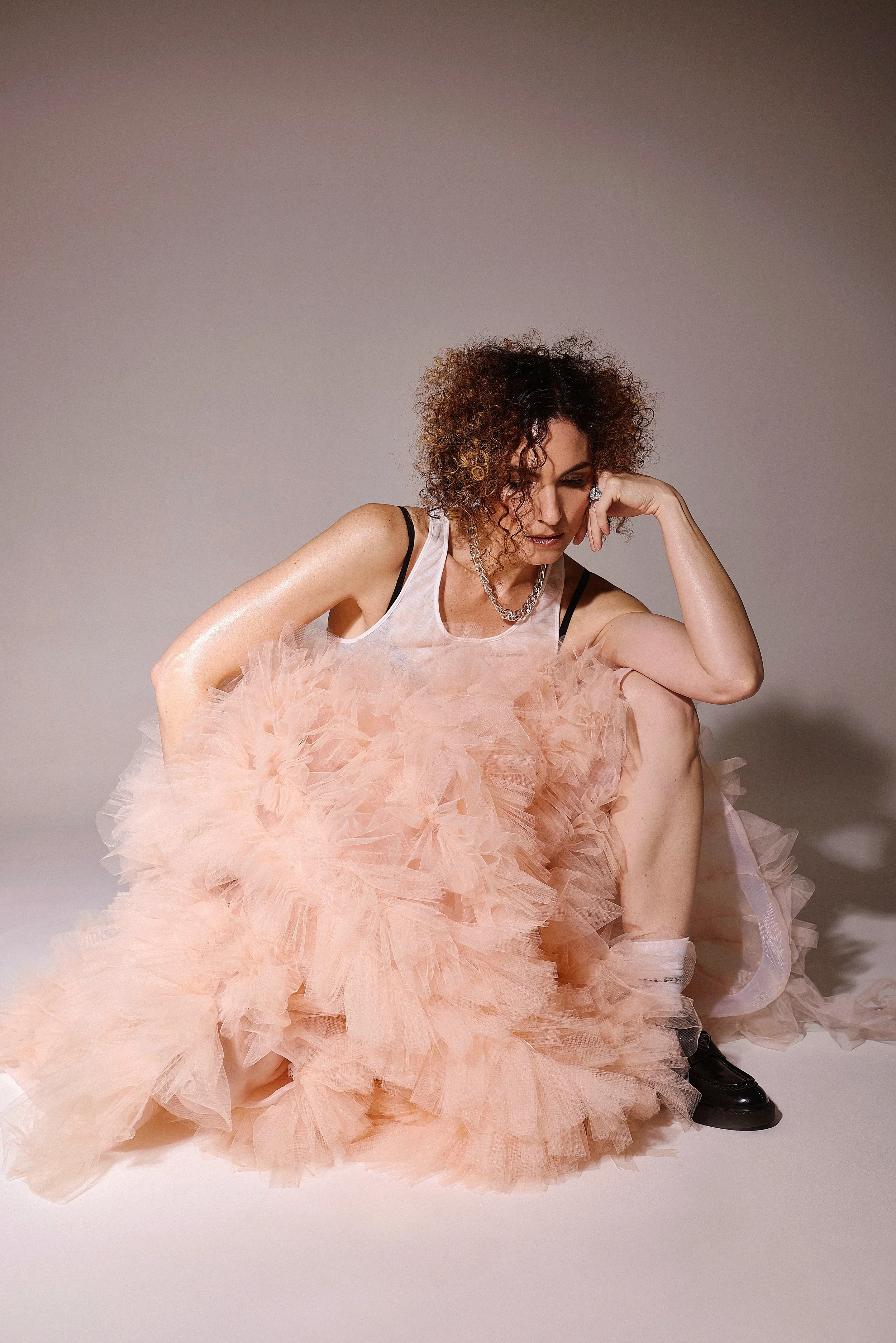 Jessica O'Donoghue with curly hair, wearing a jewelry necklace and earrings, dressed in a pink ruffled dress, is in a seated pose with her head resting on her hand, against a plain gray background.