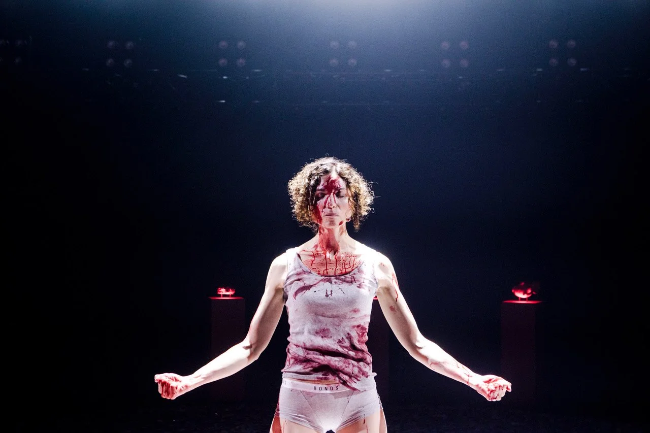 Jessica O'Donoghue with curly hair, covered in fake blood, standing in a dark room with stage lights. She is wearing a bloodstained tank top and underwear, with her arms extended outward. There are two platforms with red lights behind her.