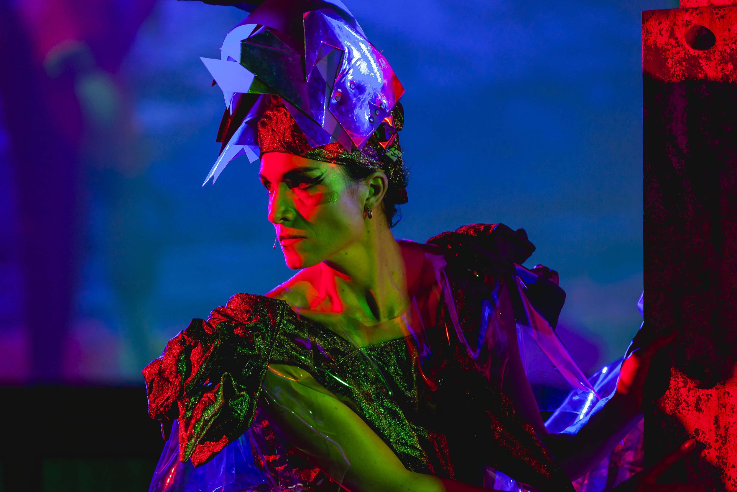 Jessica O'Donoghue wearing a colorful, metallic, and holographic outfit with a large, shiny headpiece under vibrant multicolored lighting.