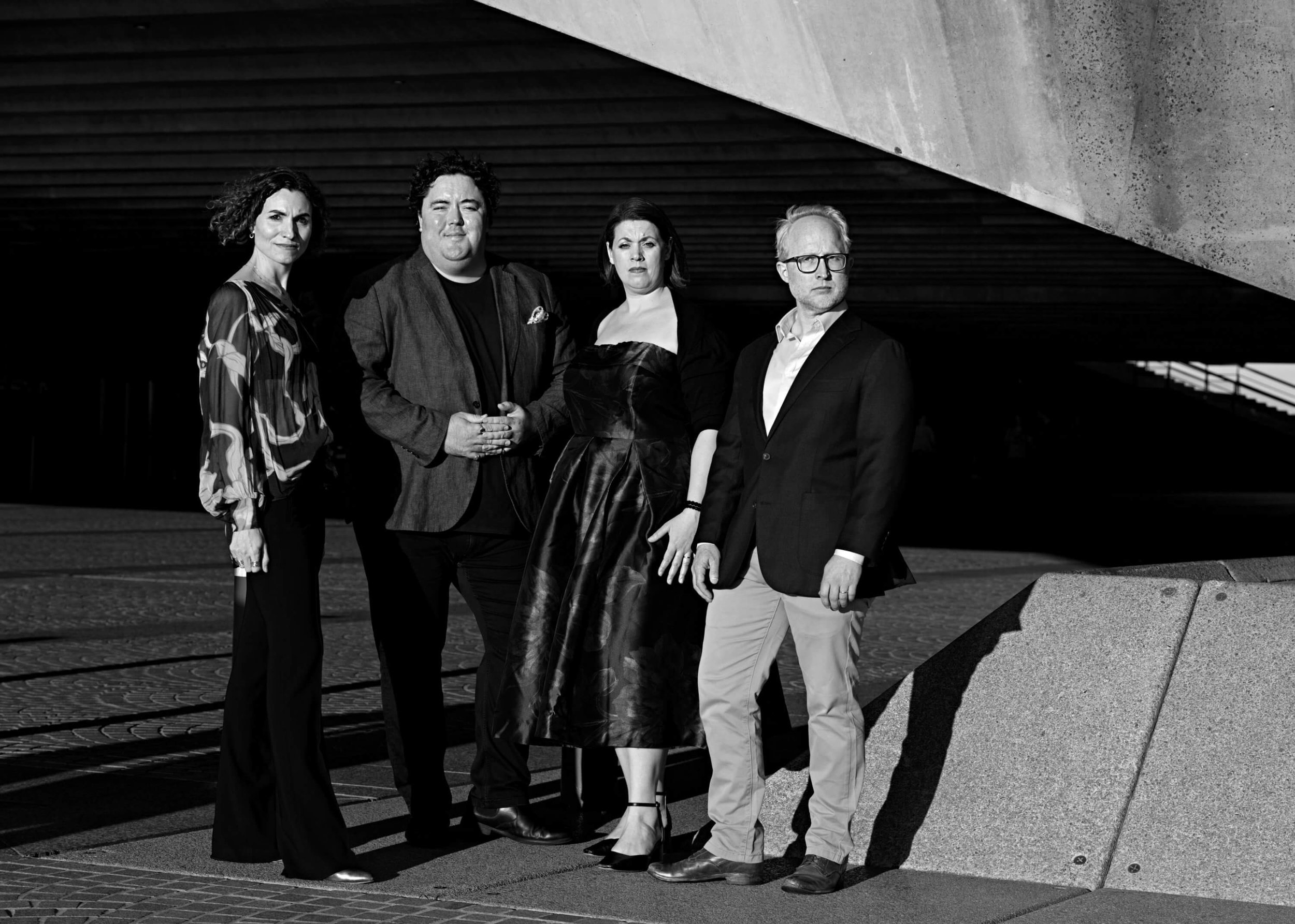 A black-and-white photo of five diverse adults standing outdoors under a modern structure with dark horizontal beams and concrete. They are dressed formally and looking at the camera.