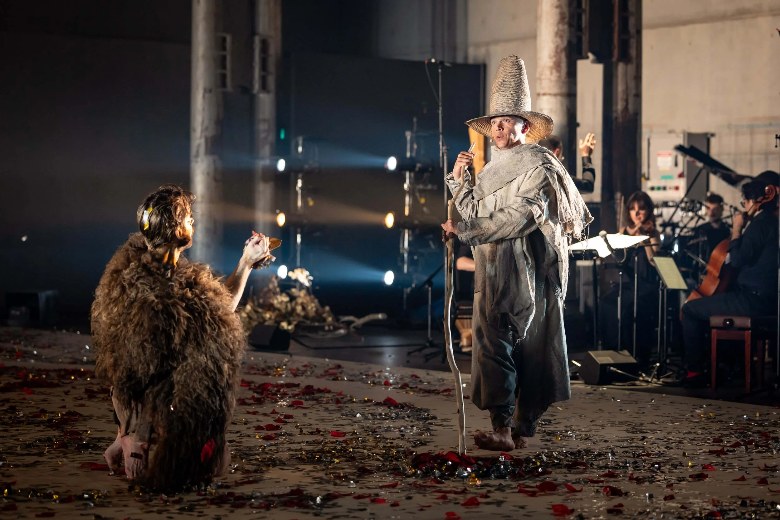 A theatrical performance featuring actors in costumes. One actor is dressed in a ragged outfit with a tall, cone-shaped hat and is holding a staff, engaging with another actor in a furry costume. Musicians are visible in the background, playing instr