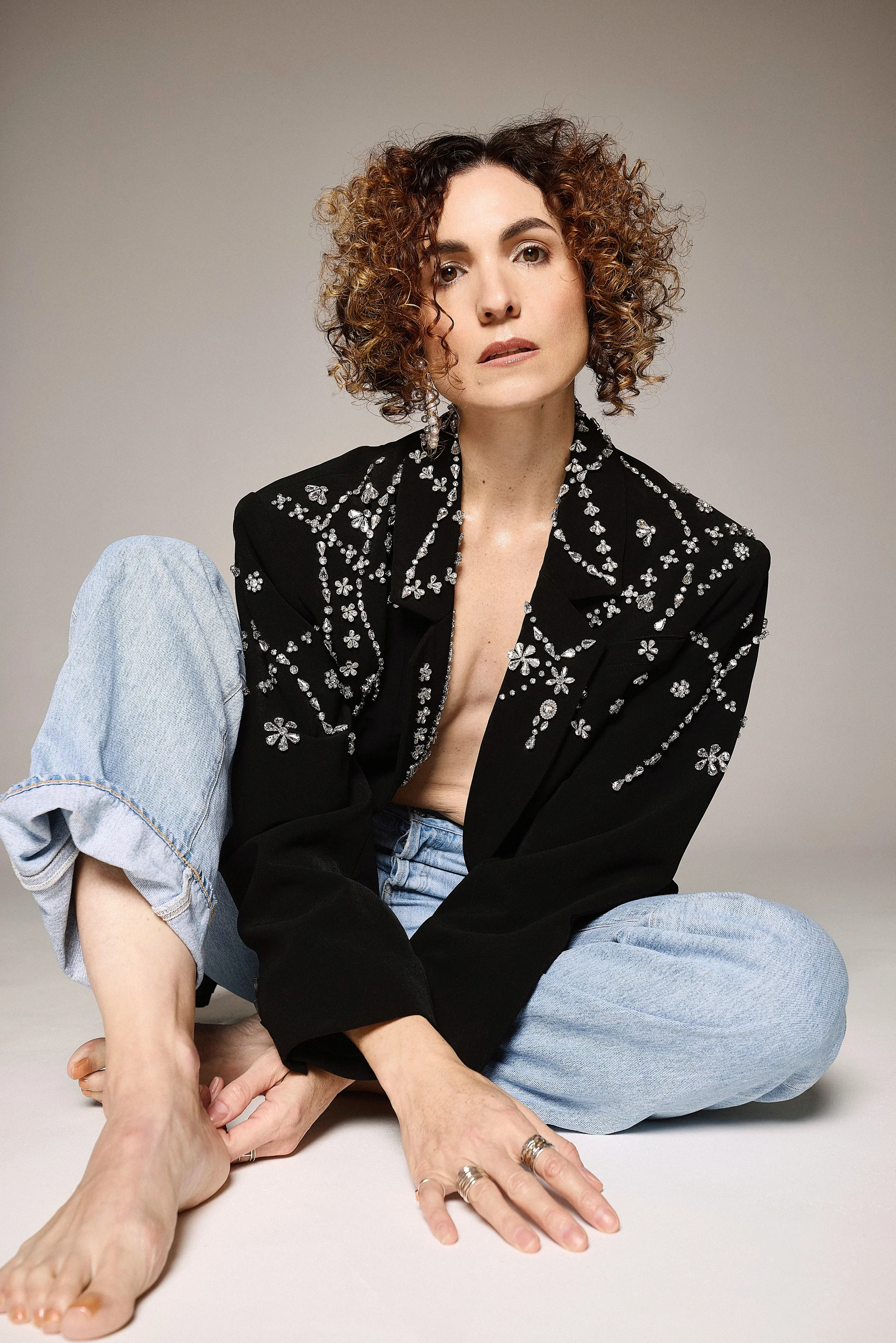 Jessica O'Donoghue with curly hair wearing a black embroidered jacket and light blue jeans sits on the floor, holding the foot of a person with a ring on their finger. The background is plain gray.