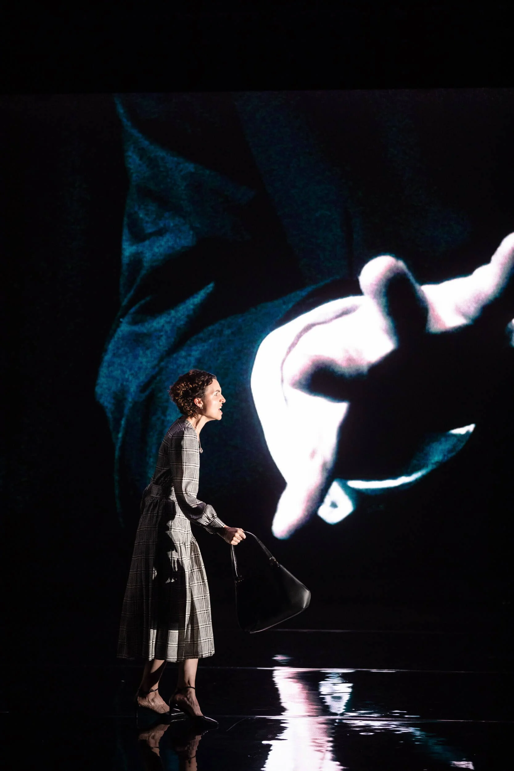 Jessica O'Donoghue in a plaid dress holding a black handbag, standing on a shiny black stage, with a large screen behind her showing an abstract, dark image with a hand reaching forward.
