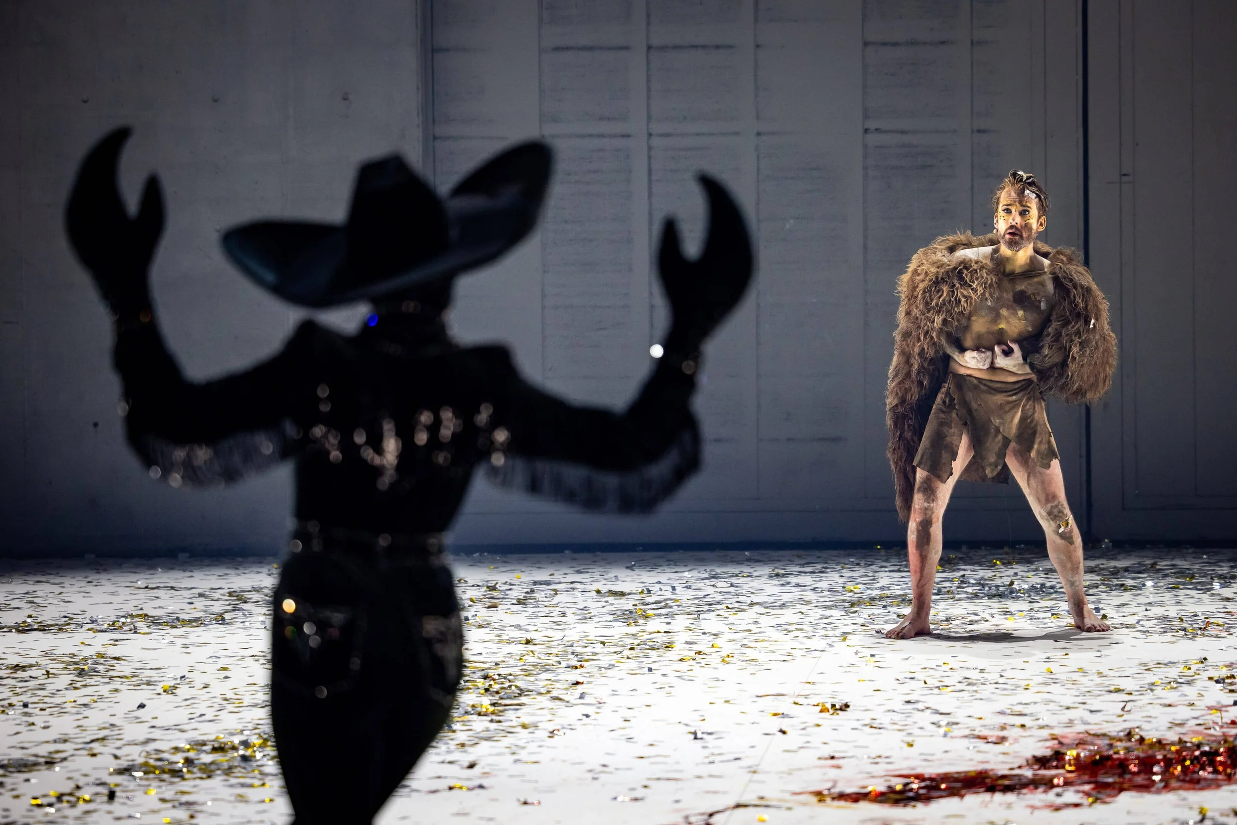 A person dressed in a costume resembling a beast or creature, standing on a stage covered in leaves and confetti, with a large black and brown prop in the foreground.