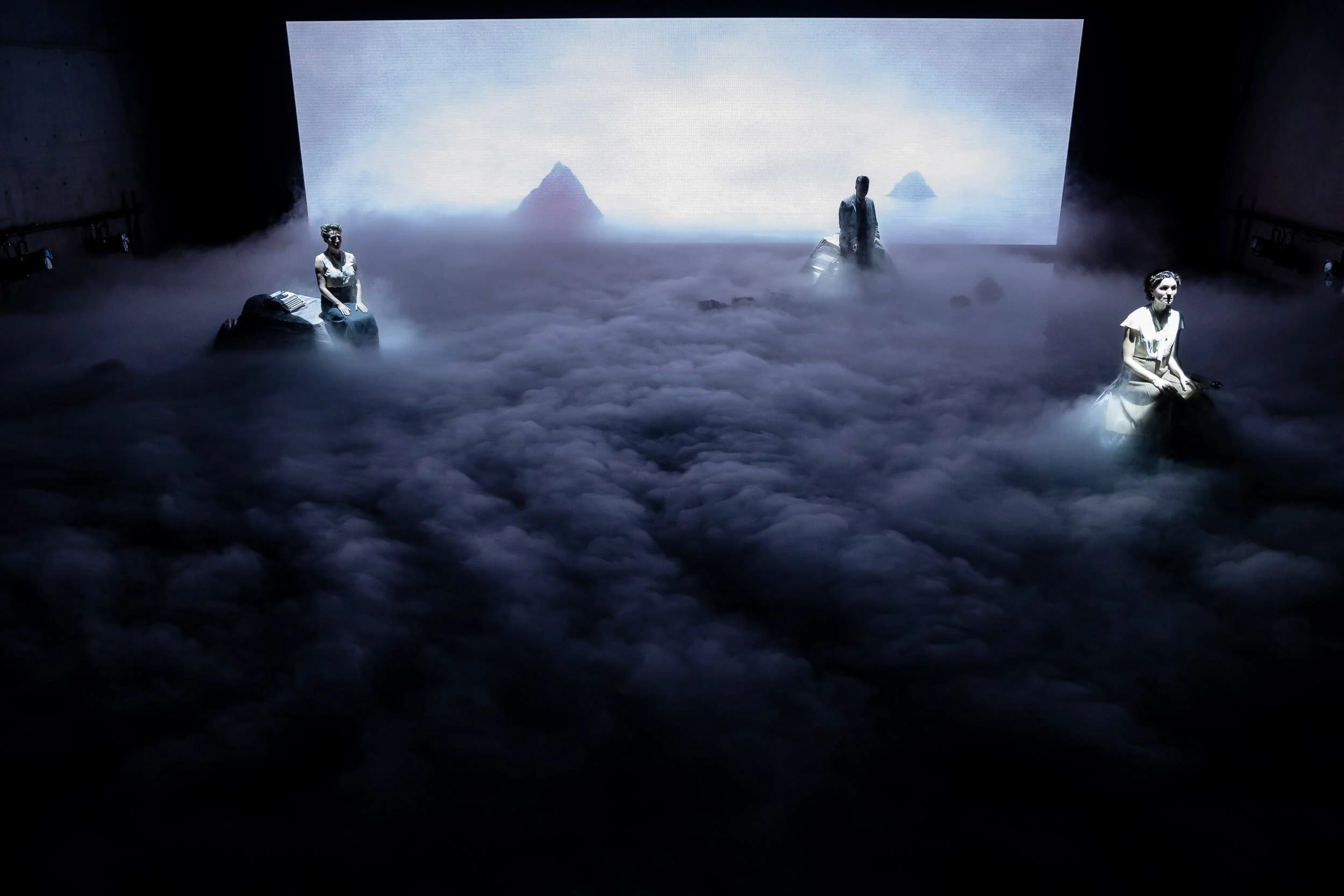 Stage scene with three performers dressed in vintage clothing, two women seated and one man standing, in front of a large backdrop displaying a sea with islands and fog, creating an atmospheric setting.