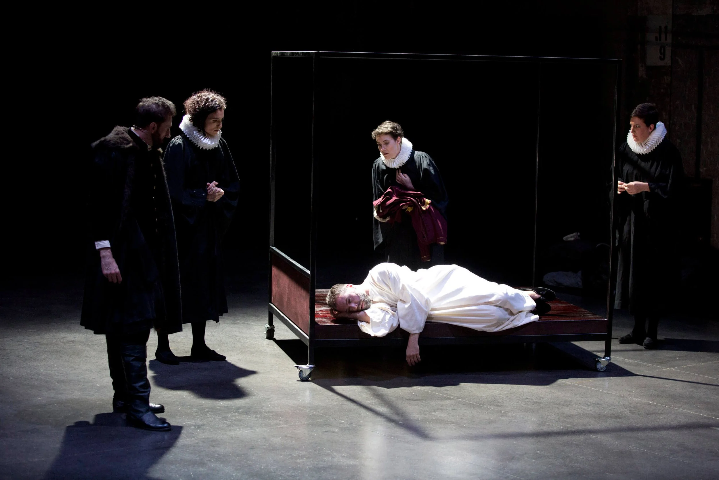 The image depicts a theatrical stage scene with five actors dressed in historical costumes. Four actors are standing around a stretcher on which a person is lying, appearing to be unconscious or deceased. The scene is dark, with focused lighting on t
