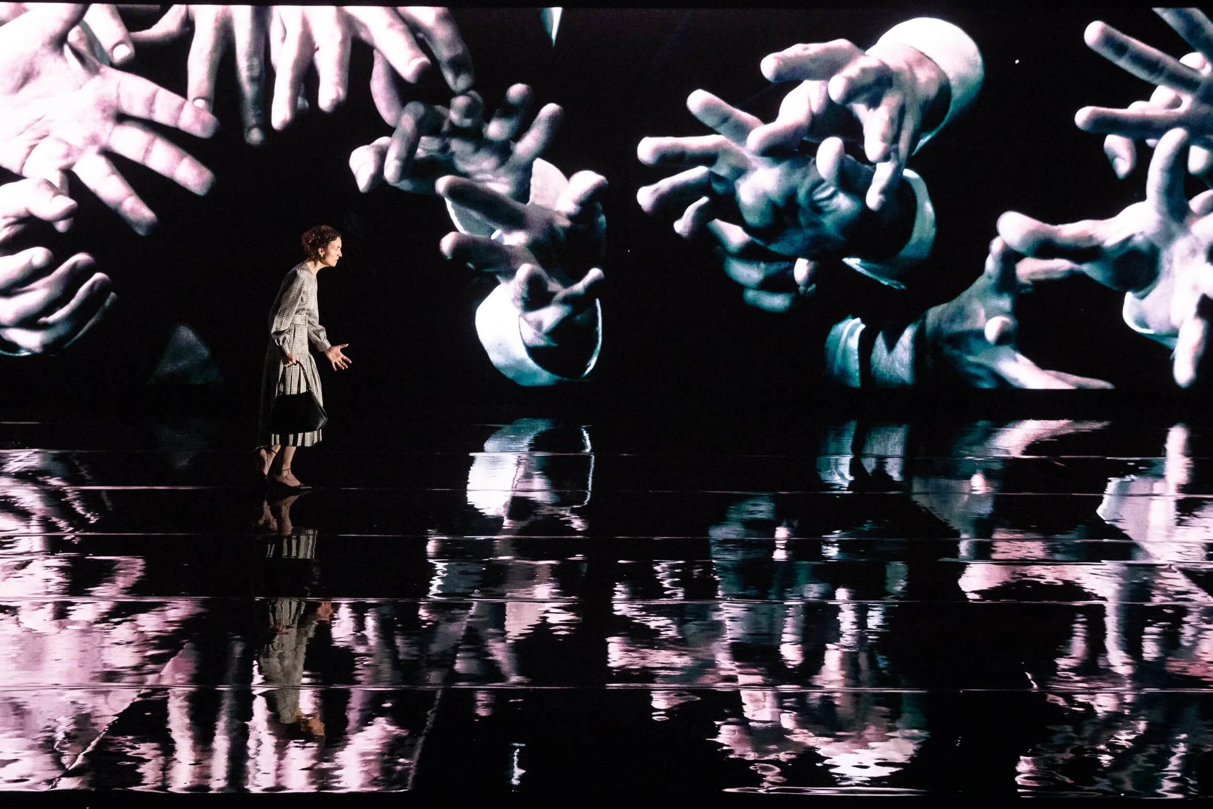 Jessica O'Donoghue stands on a stage in front of a large digital display showing multiple hands reaching in various directions. The reflection of the scene is visible on the shiny floor.