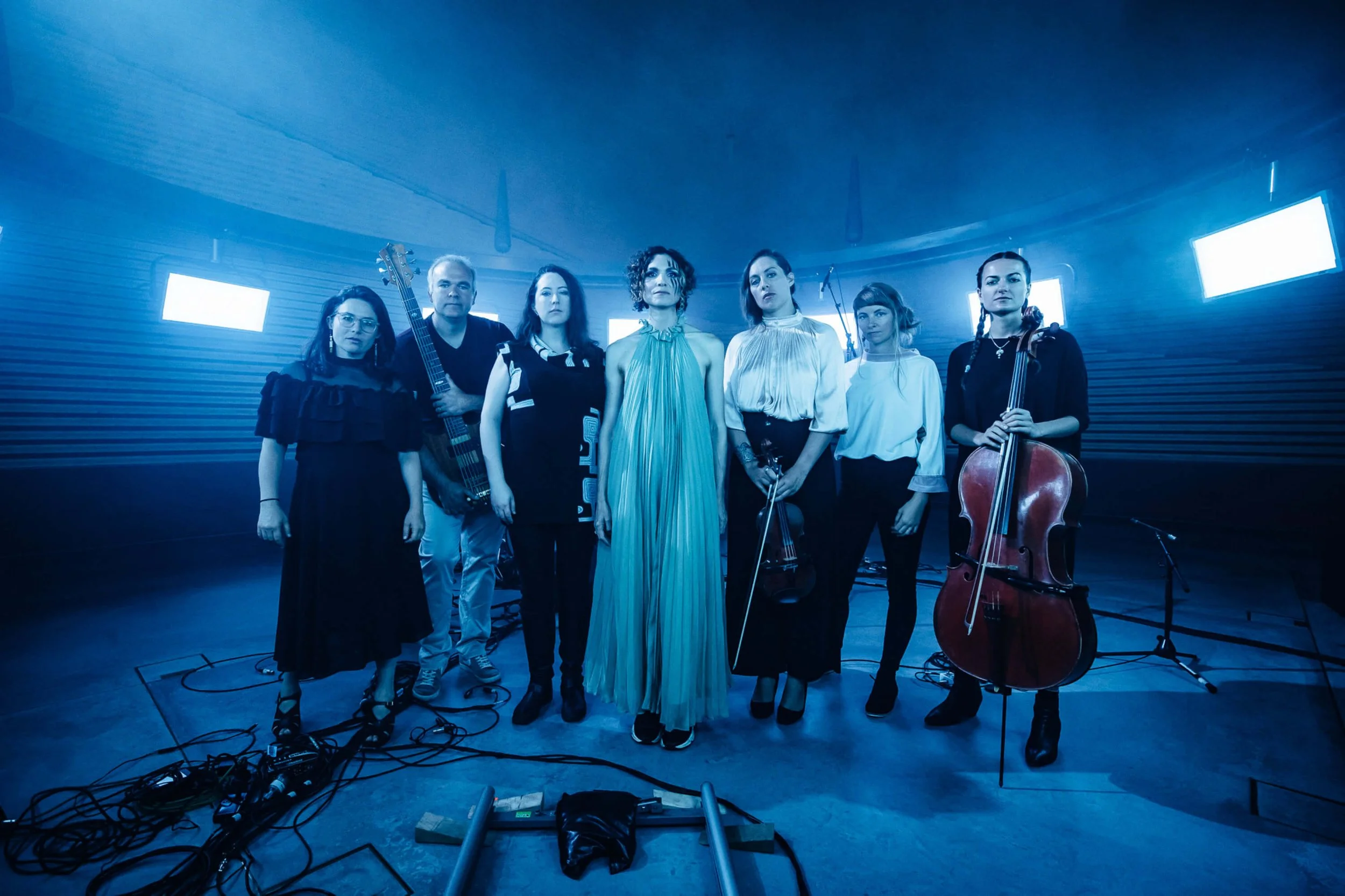 Group of six women and one man standing in a music studio, holding musical instruments including a guitar, violin, and cello, with blue lighting and microphones in the background.